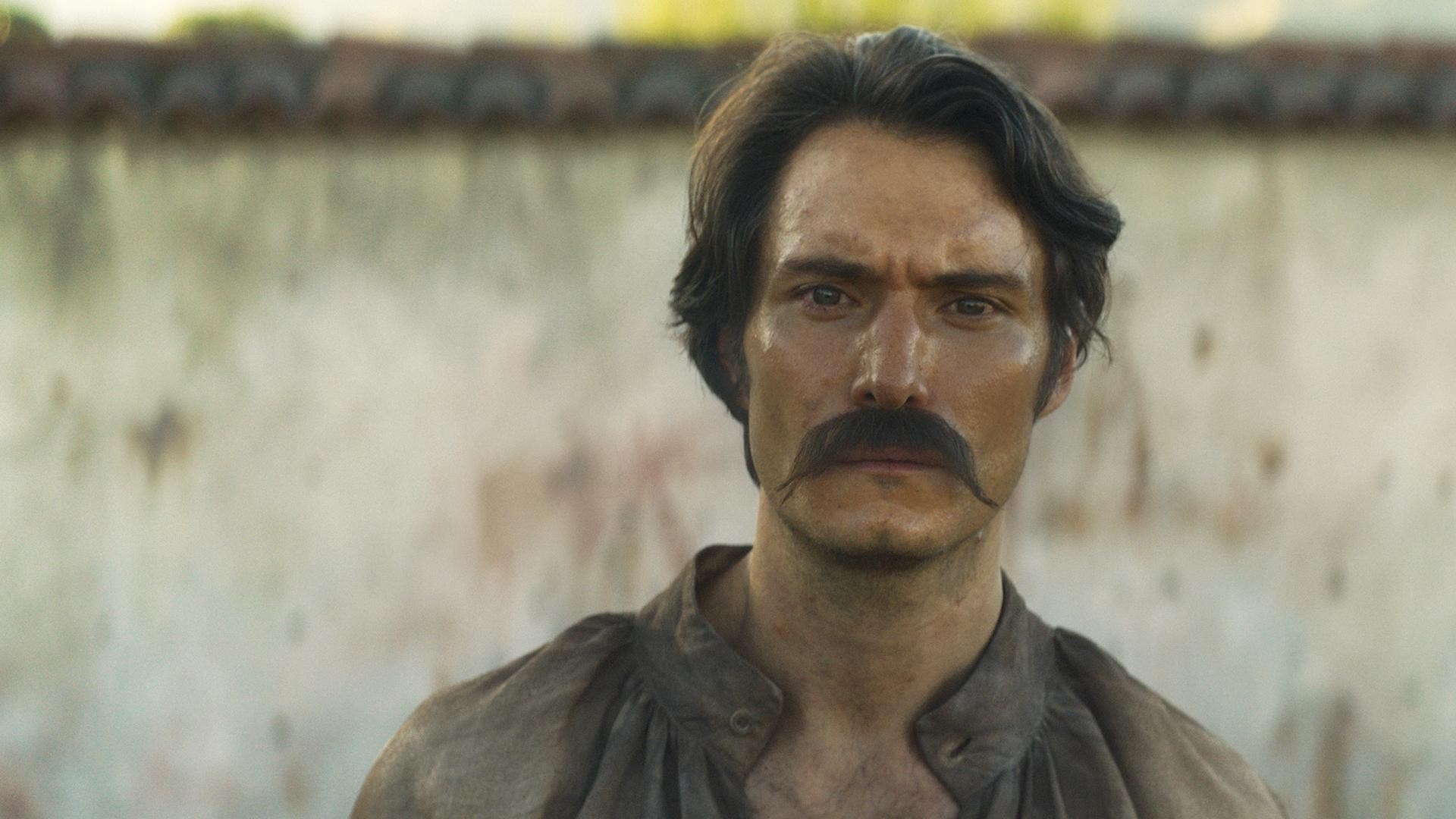 Claudio Cataño en primer plano como el coronel Aureliano Buendía, con bigote y mirada fija, frente a un muro desgastado