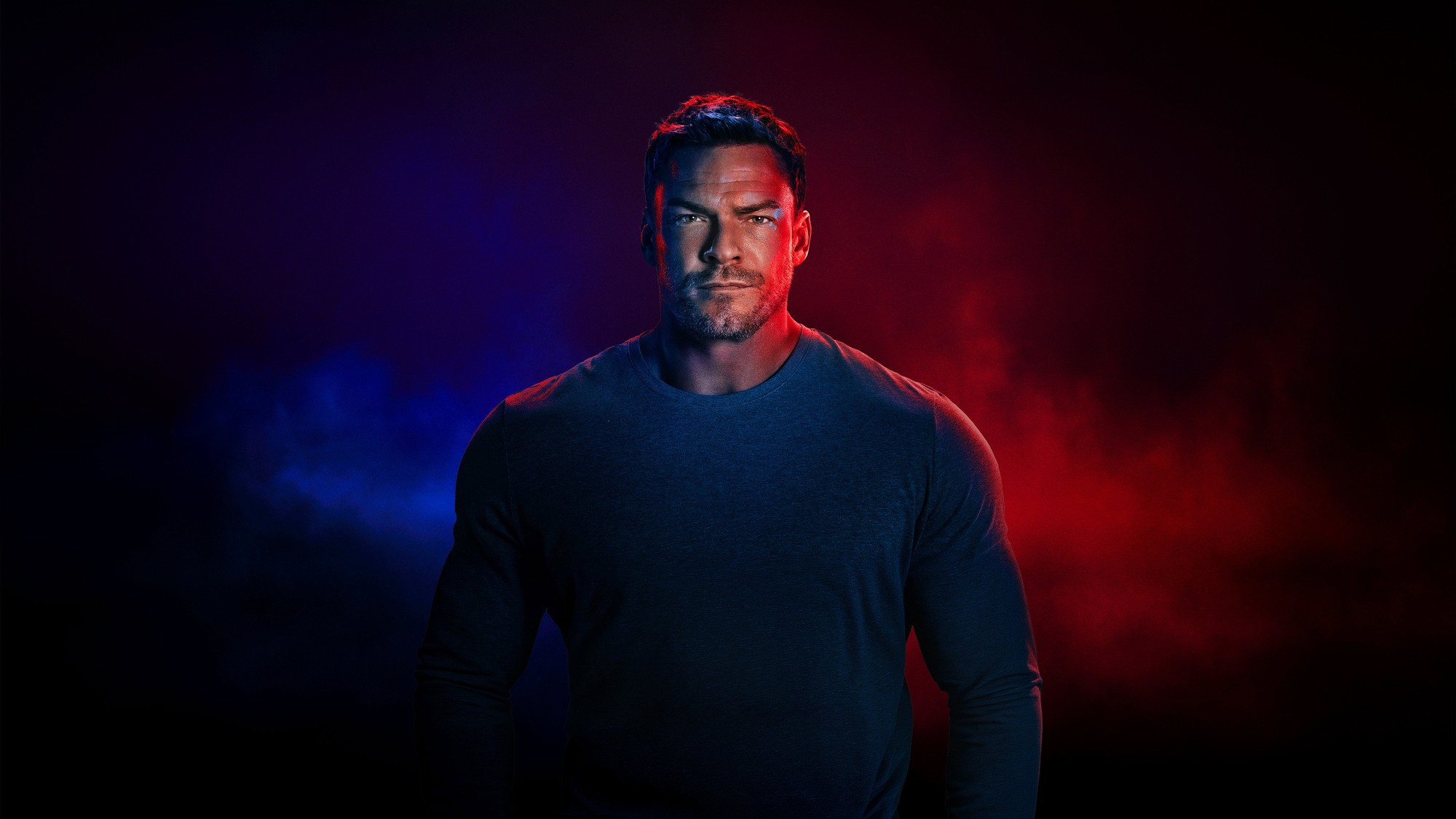 Alan Ritchson caracterizado como Jack Reacher, de pie y con la mirada fija sobre un fondo rojo y azul