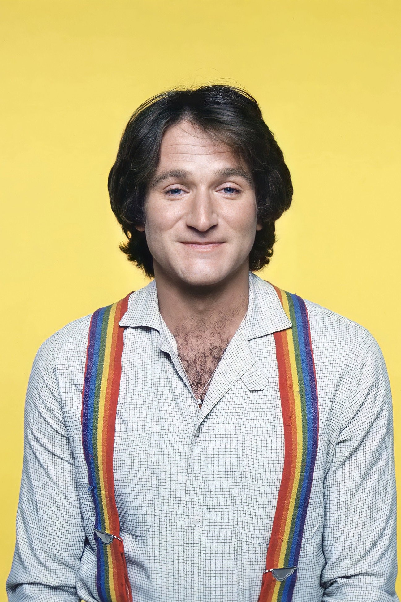 Retrato de prensa de Robin Williams joven, sonriendo con camisa clara y tirantes de colores sobre fondo amarillo
