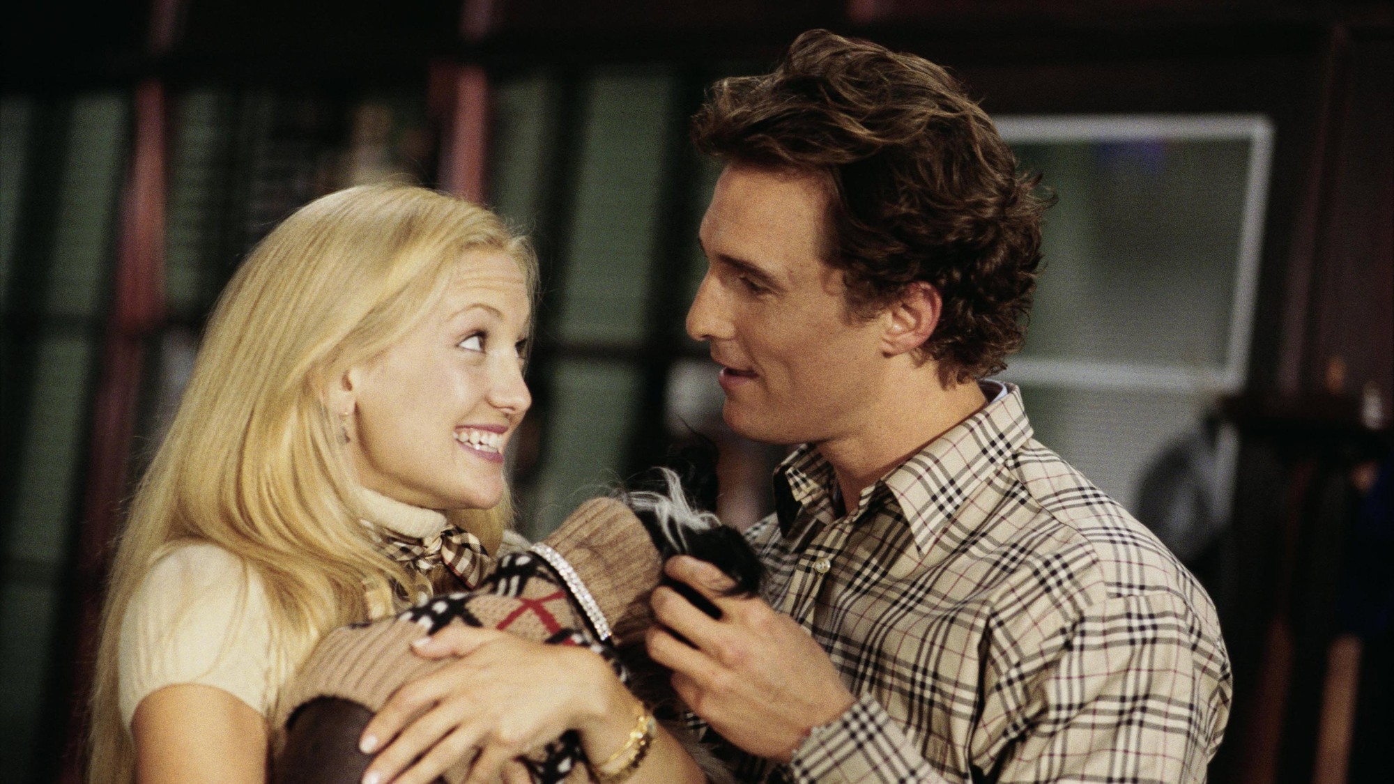 Kate Hudson y Matthew McConaughey se miran de cerca mientras sostienen juntos a un perro pequeño dentro de un departamento