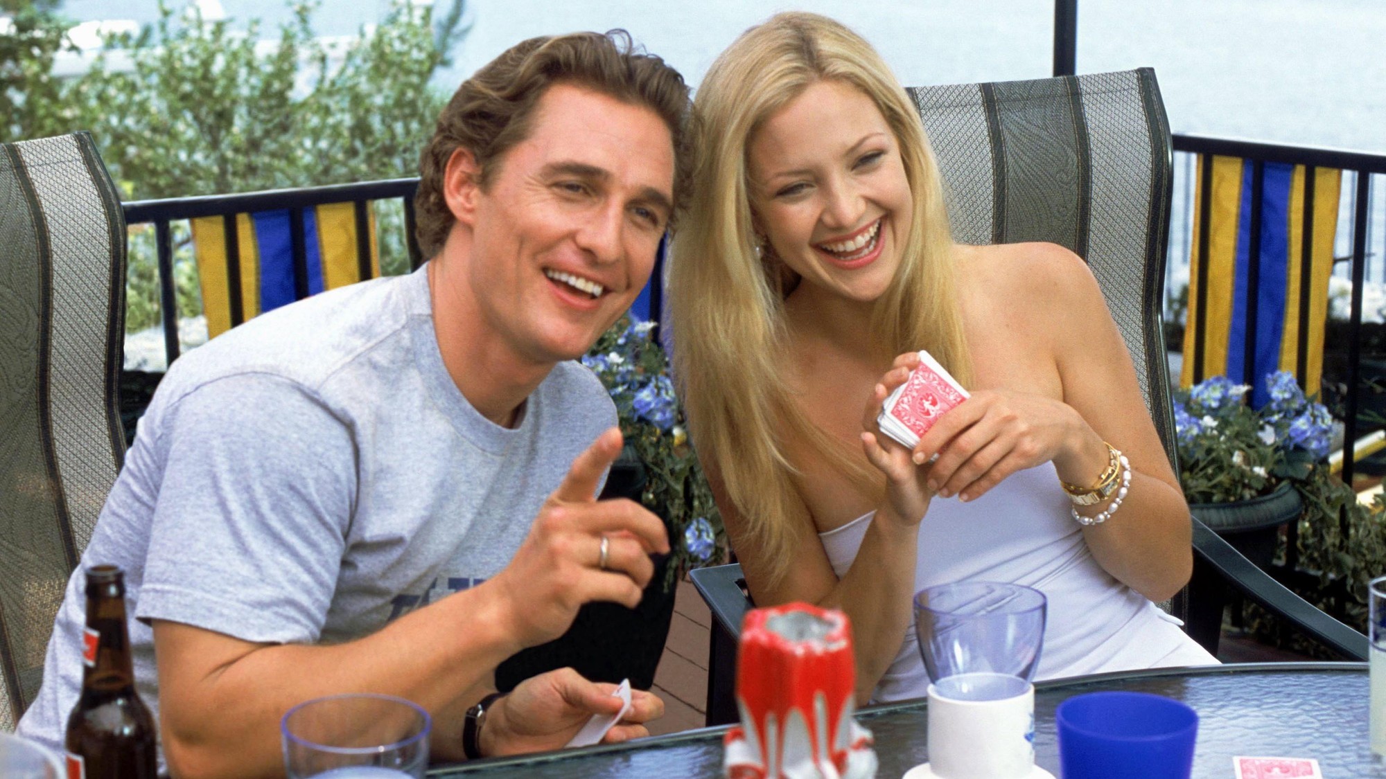 La pareja protagonista conversa y ríe alrededor de una mesa de jardín durante una reunión familiar al aire libre