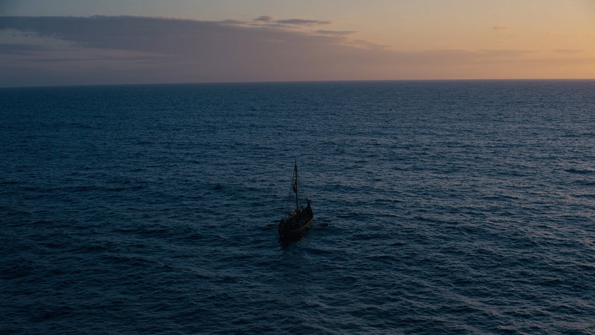 Un barco solitario navega en mar abierto bajo un cielo de atardecer en una escena de The Odyssey