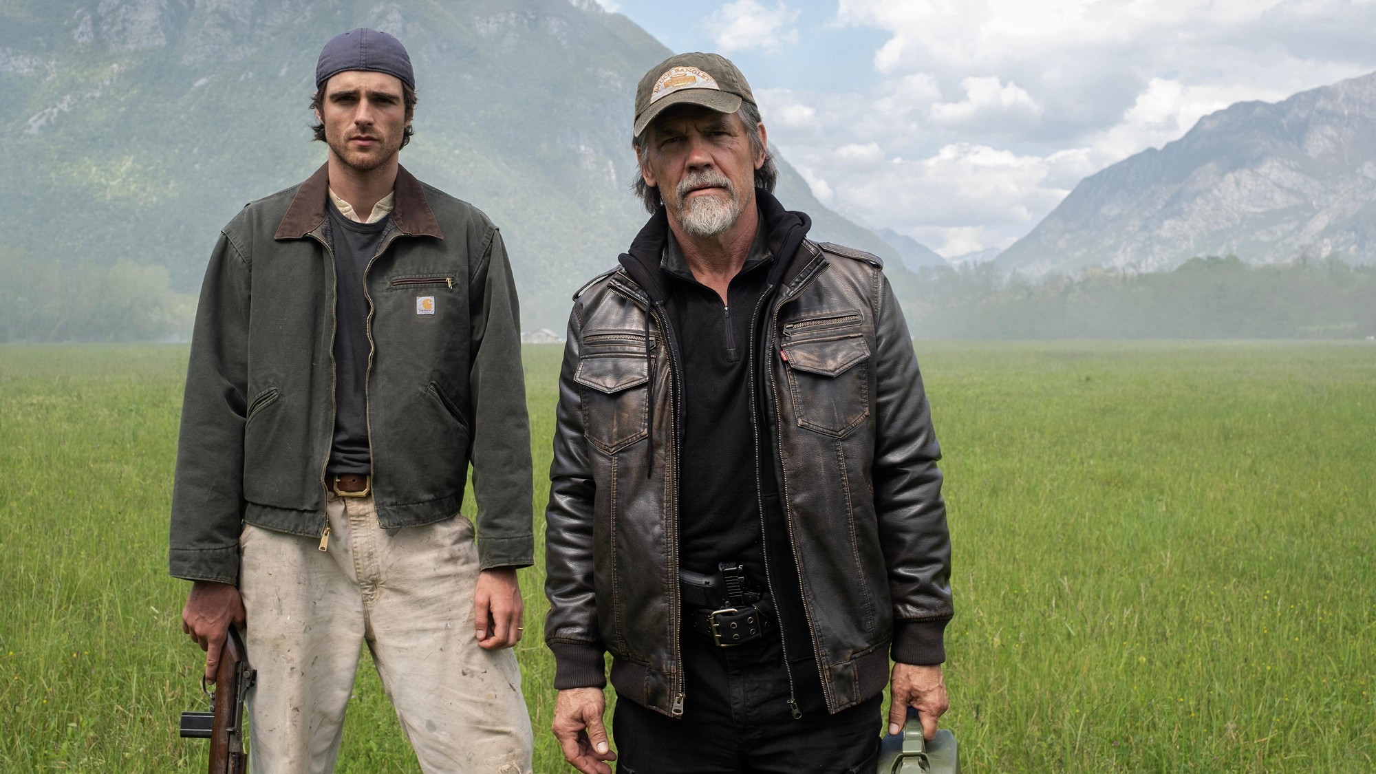 Jacob Elordi con un rifle y Josh Brolin con un bidón de combustible, de pie en un prado verde ante montañas, en The Dog Stars