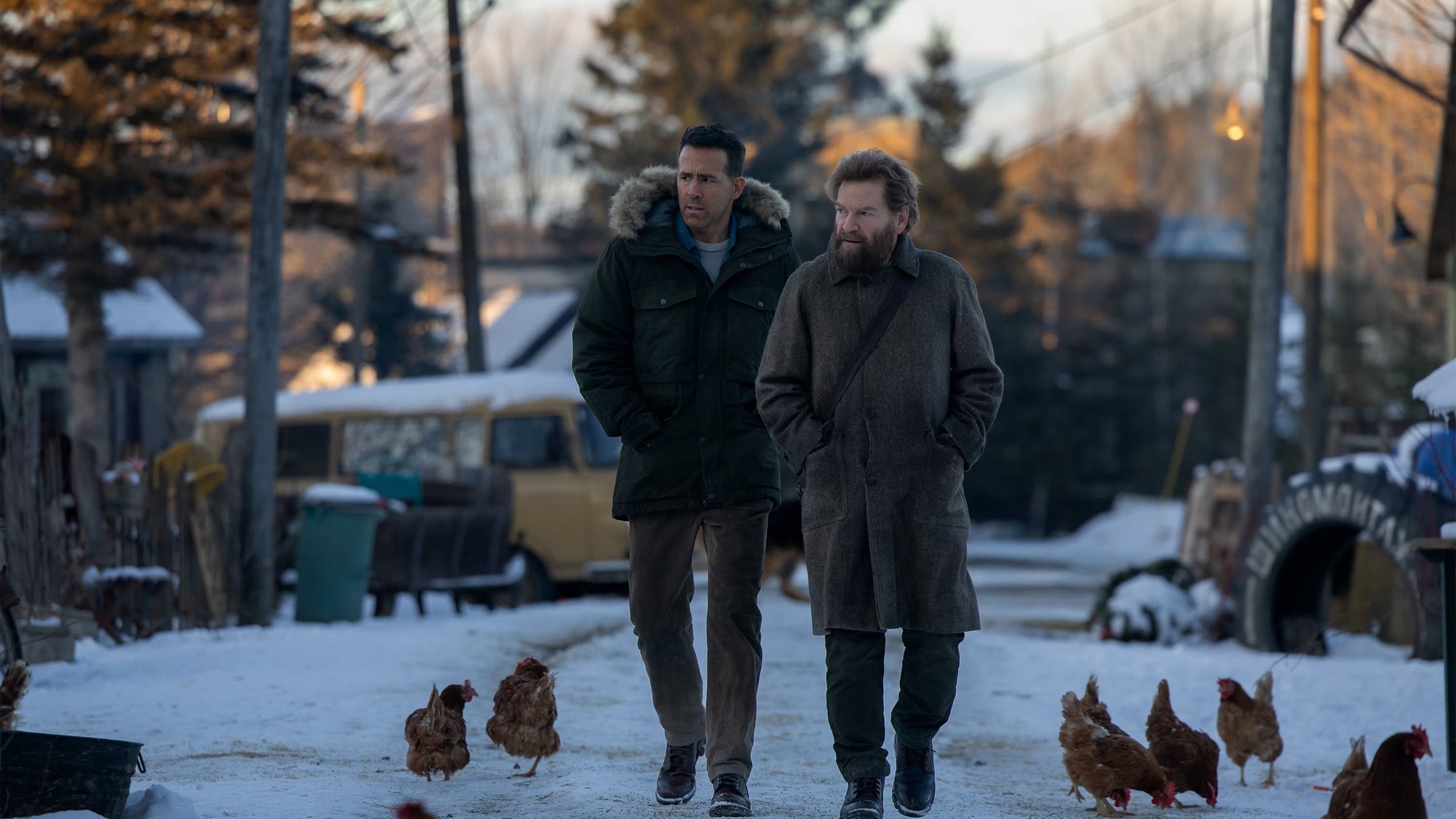 Ryan Reynolds y Kenneth Branagh caminan por un camino nevado de una aldea rusa al atardecer, rodeados de gallinas y camionetas viejas