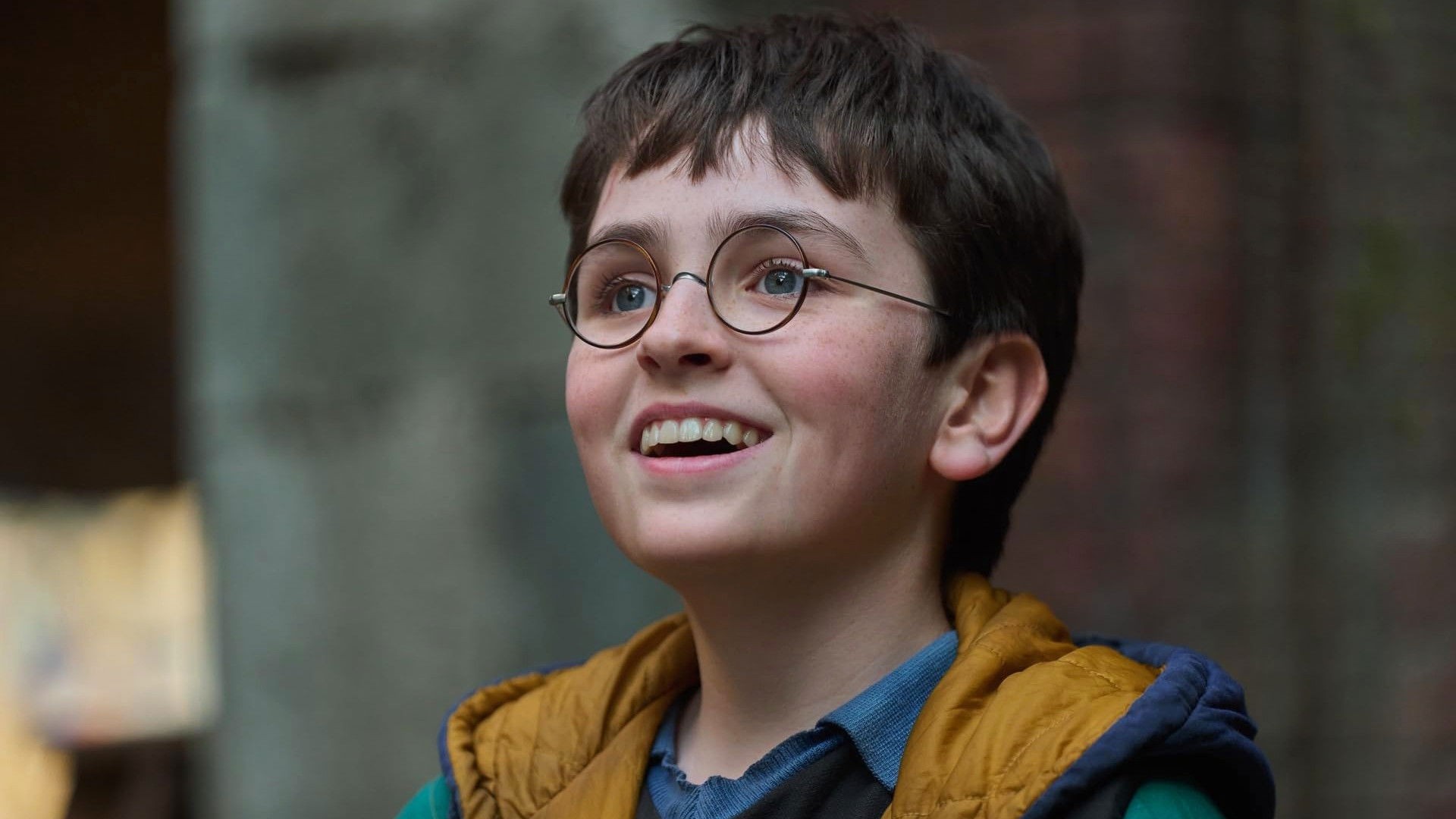 Dominic McLaughlin sonríe caracterizado como Harry Potter, con sus gafas redondas y un abrigo, en la serie de HBO Max