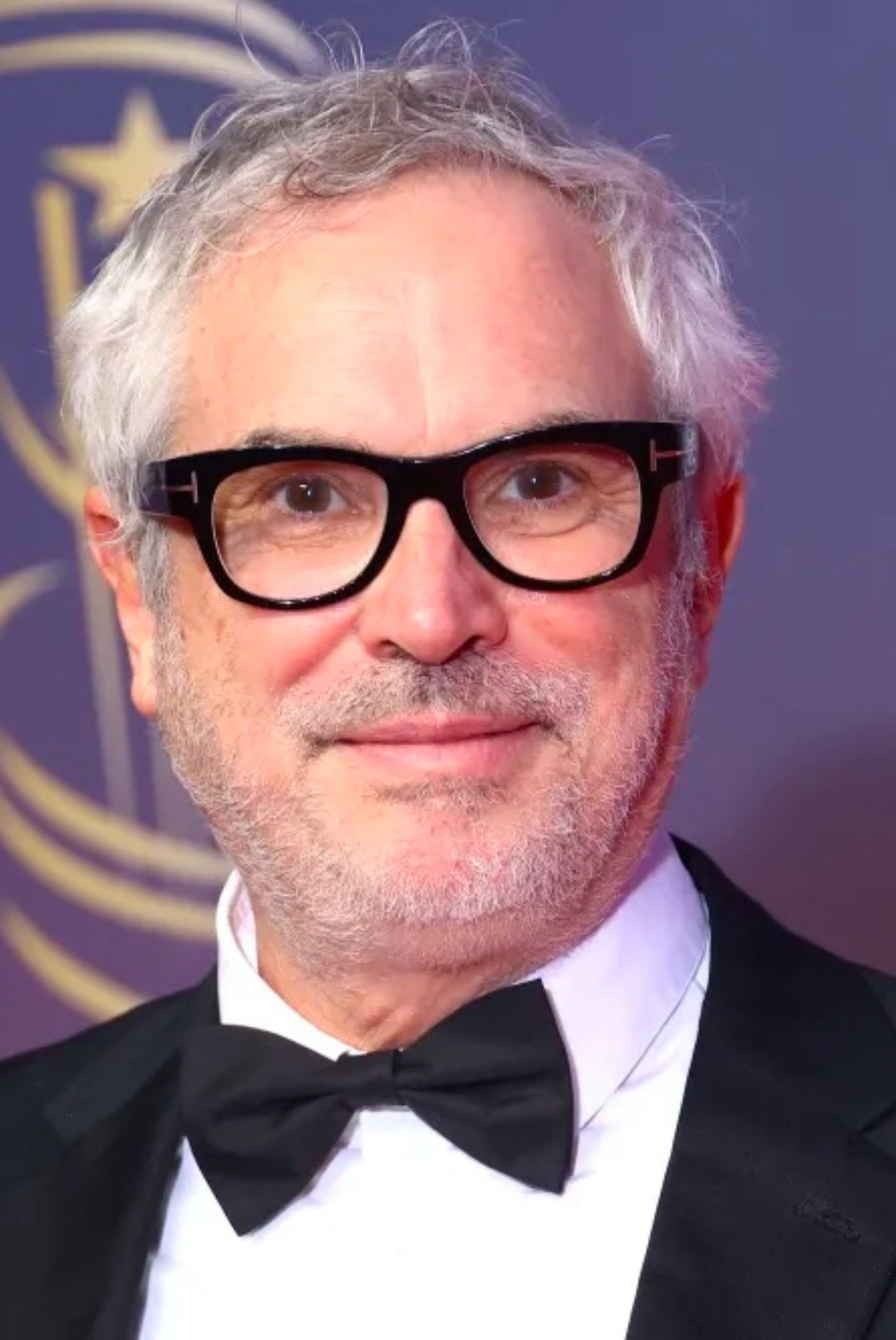 Retrato del cineasta mexicano Alfonso Cuarón con lentes de armazón oscuro y esmoquin durante una alfombra roja