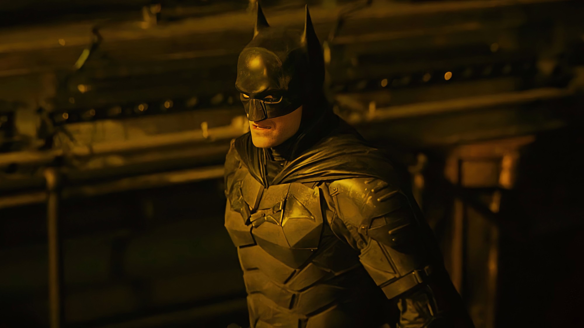 El traje completo de Batman iluminado por una luz ámbar dentro de un espacio industrial en penumbra