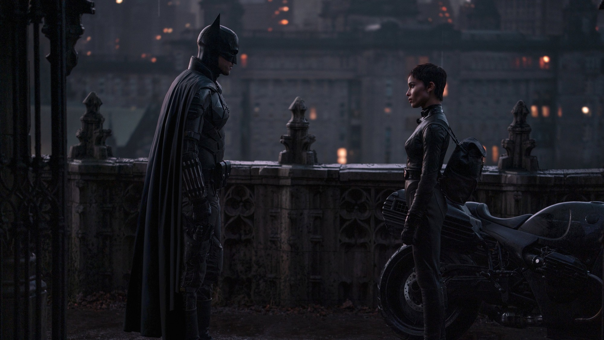 Batman y Selina Kyle frente a frente en una azotea de Ciudad Gótica, junto a una motocicleta y con la ciudad iluminada al fondo