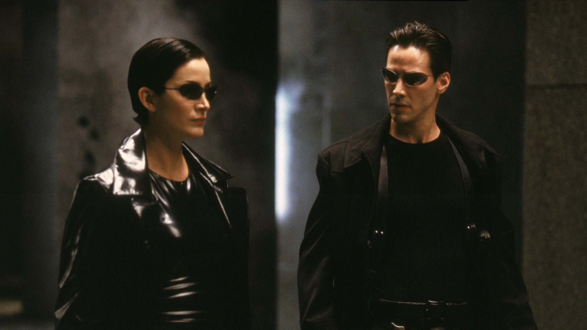 Trinity y Neo, con lentes oscuros y abrigos negros, avanzan juntos por un interior en penumbra