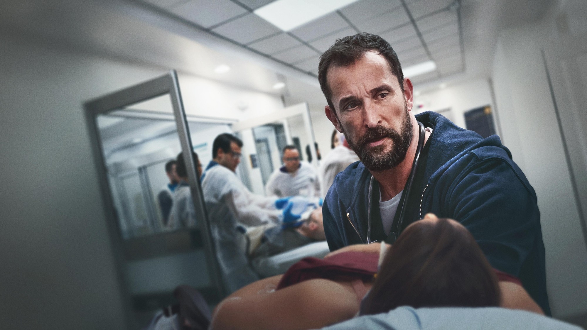 Médico con barba y sudadera azul atiende a una paciente en el pasillo de una sala de urgencias mientras el equipo trabaja al fondo