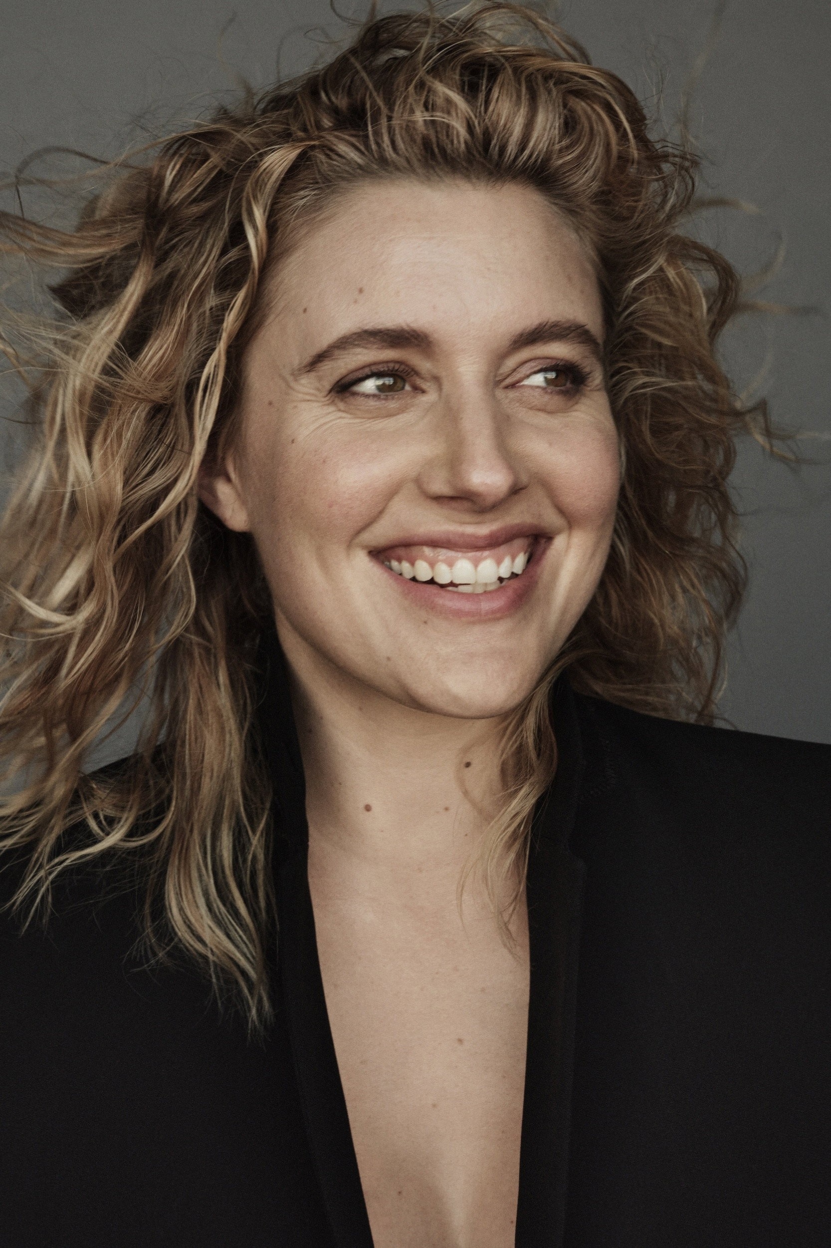 La cineasta Greta Gerwig sonriendo de perfil con el cabello suelto sobre un fondo gris neutro