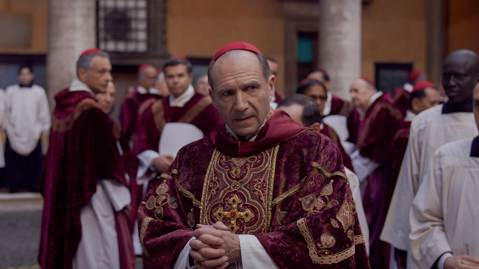 Un cardenal de vestiduras granate y dorado permanece de pie, con las manos entrelazadas, entre otros religiosos en un patio