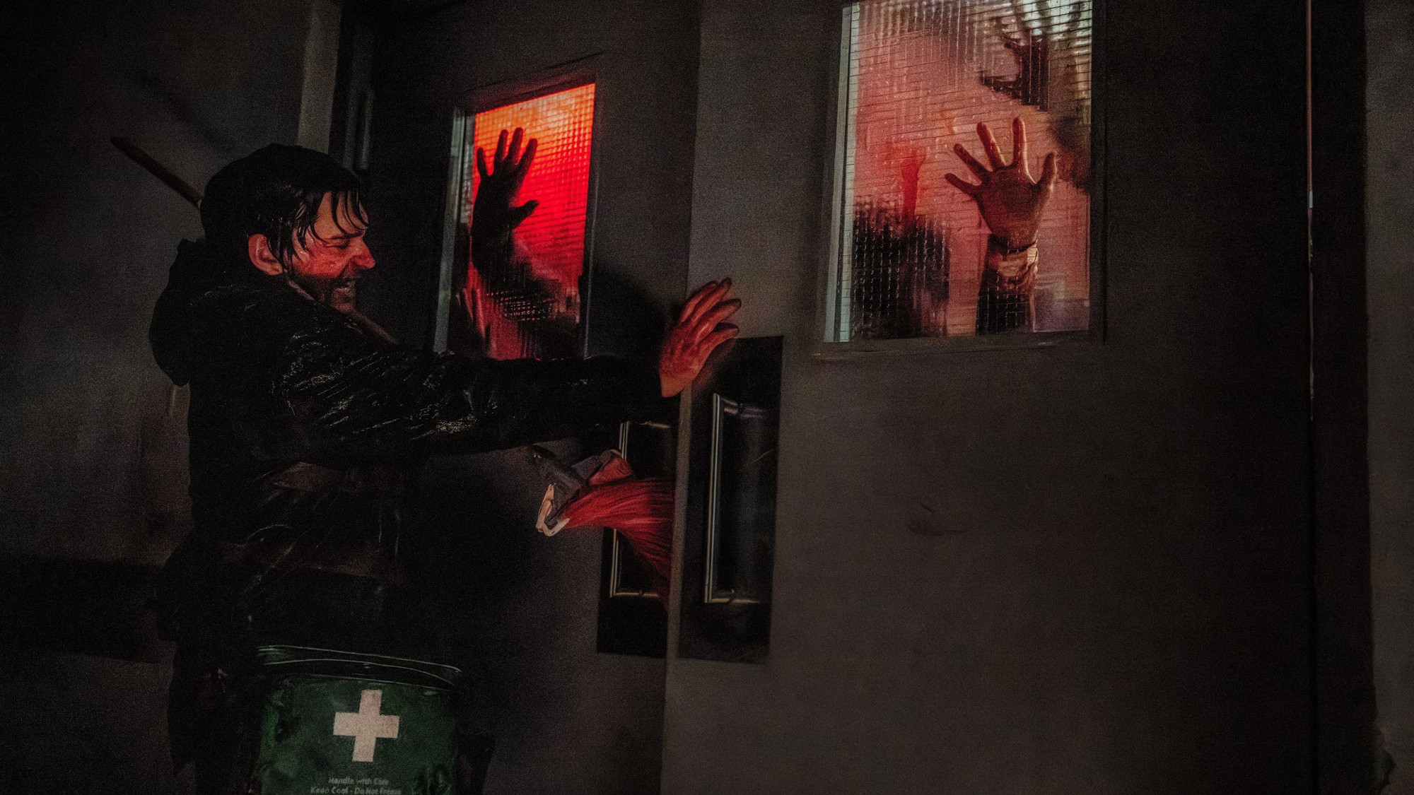 Manos de infectados presionadas contra unas ventanas iluminadas de rojo mientras un personaje bloquea la puerta desde el otro lado