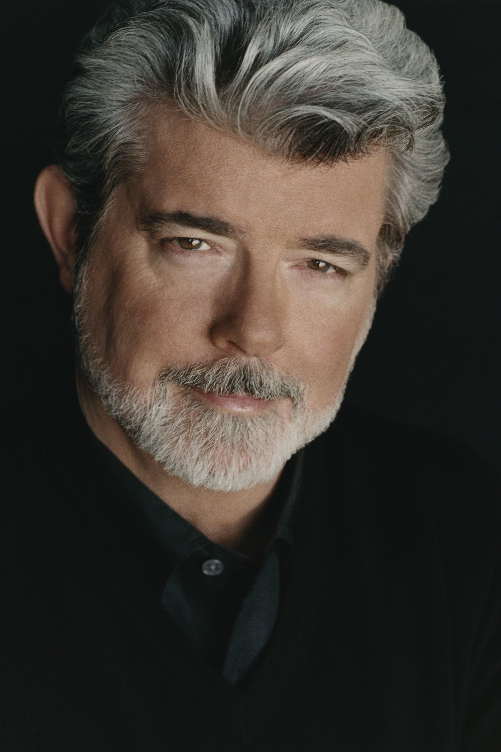 Retrato de estudio de George Lucas, de cabello canoso y barba, vestido de negro sobre un fondo oscuro