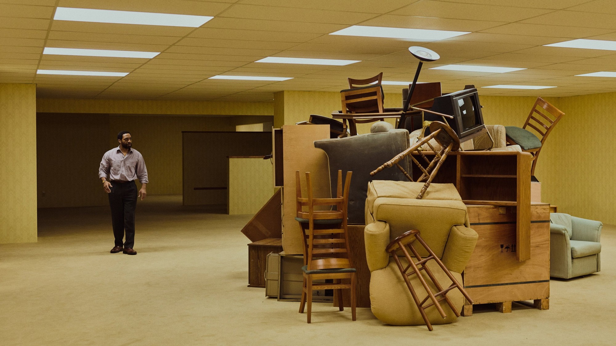 Un hombre solo de pie en una sala de exhibición de muebles con paredes amarillas, frente a una montaña de sillones y sillas apiladas