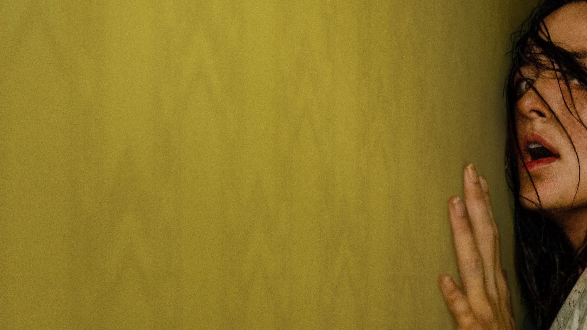 Primer plano lateral de una mujer con el cabello mojado pegado al rostro, apoyando la mano contra una pared amarilla mientras grita