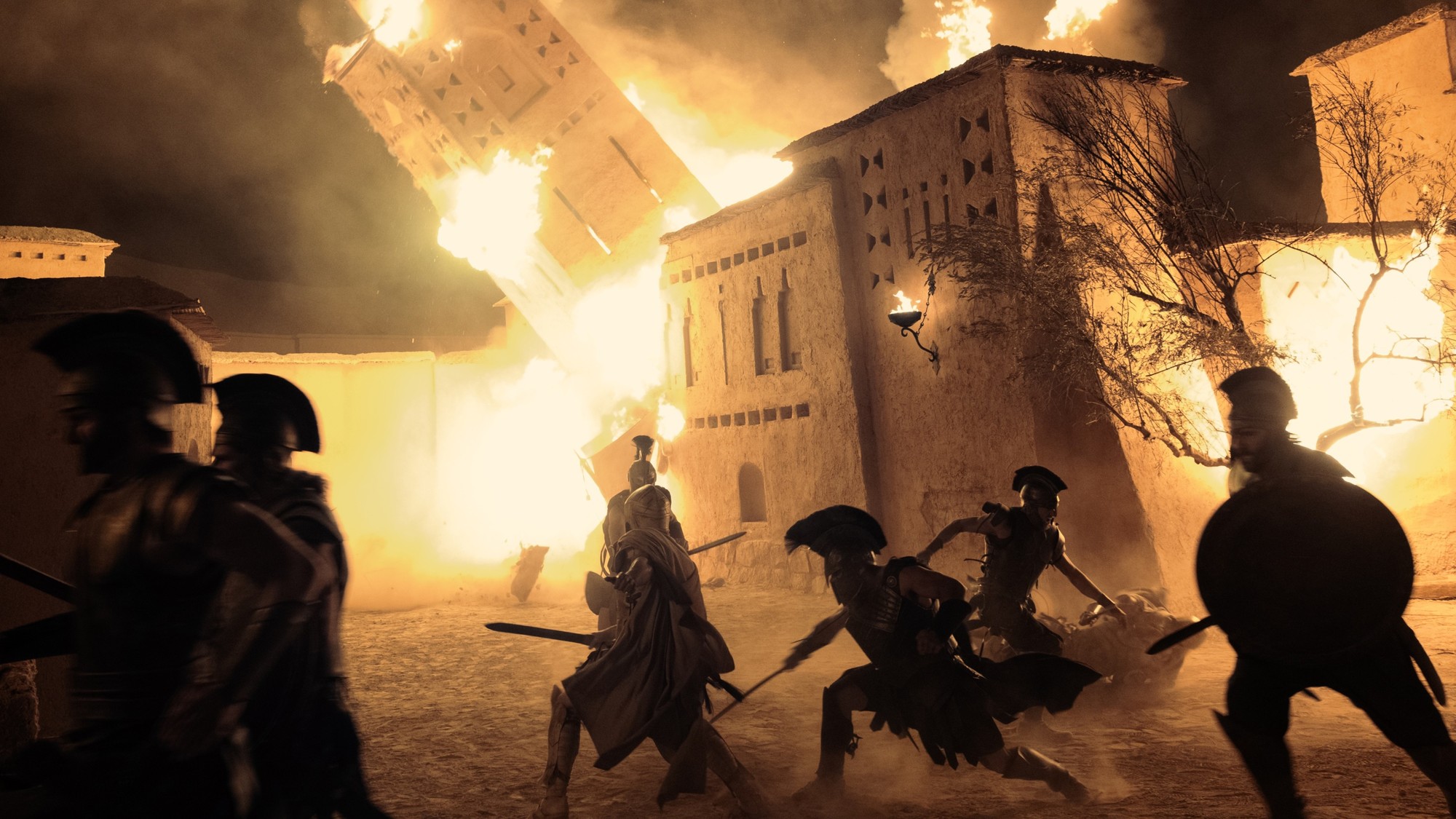 Soldados con lanzas y escudos avanzan de noche frente a una fortaleza en llamas en una escena de The Odyssey de Christopher Nolan
