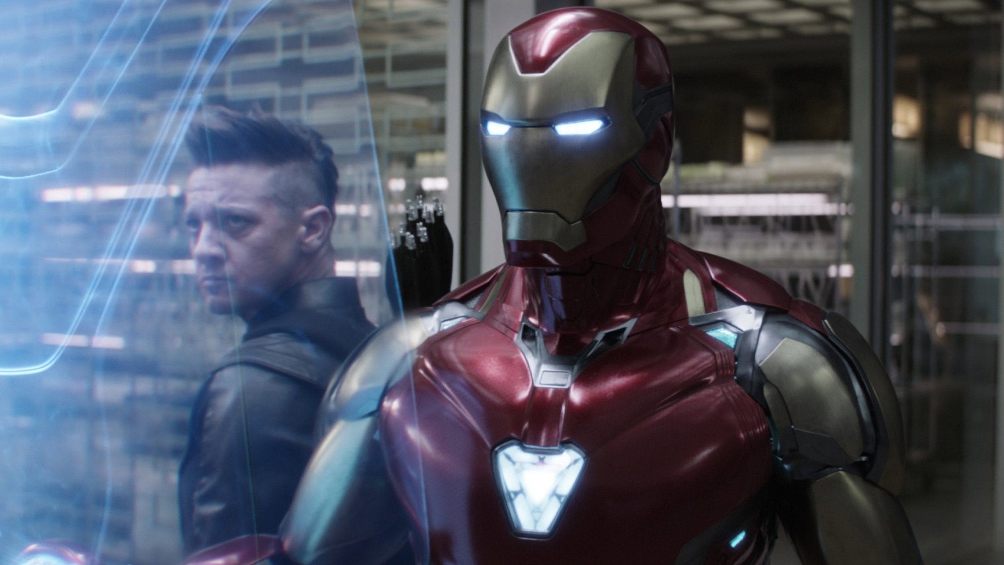 Iron Man con su armadura roja y dorada encendida junto a Ojo de Halcón en un pasillo iluminado del complejo de los Vengadores