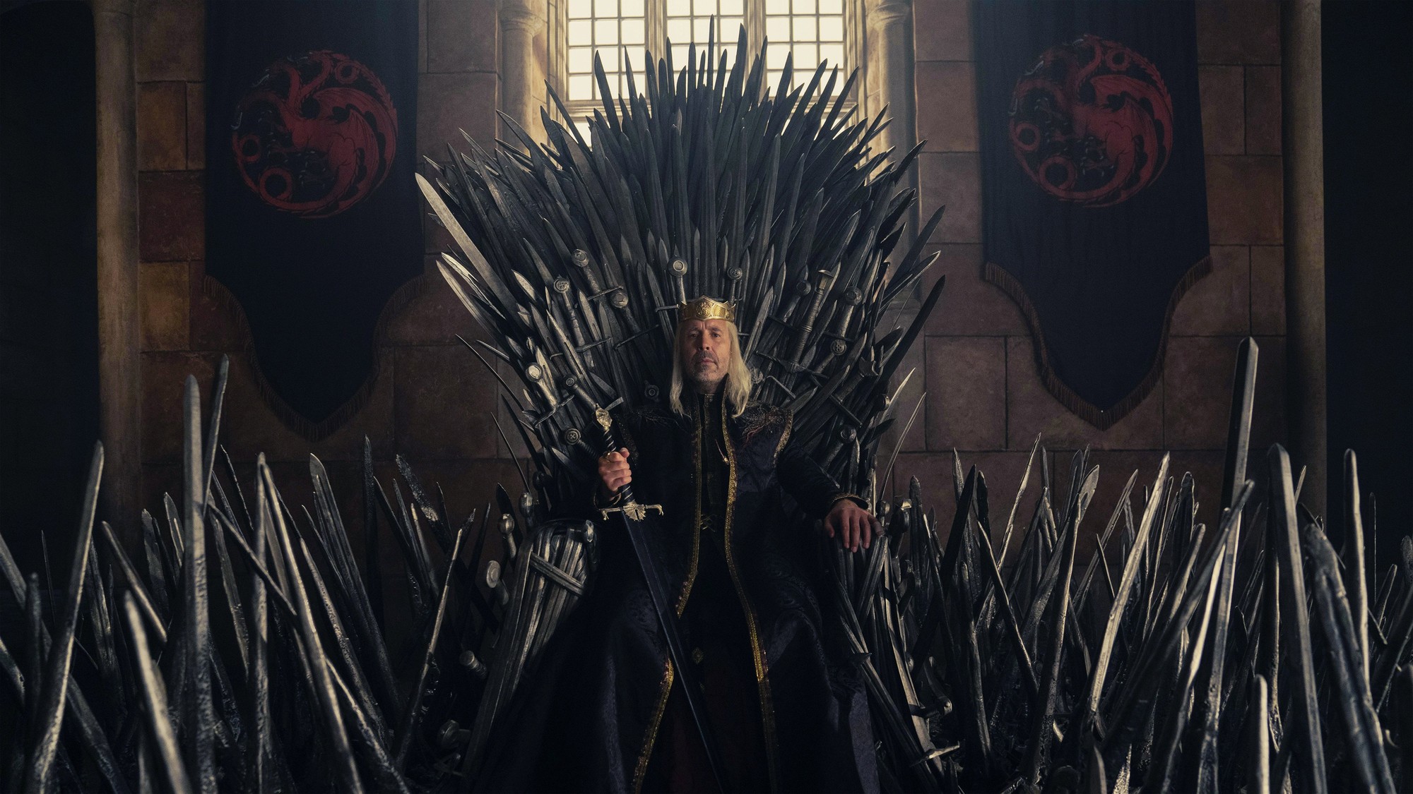 Rey anciano con corona empuñando una espada sobre el Trono de Hierro, entre estandartes rojos de la casa Targaryen
