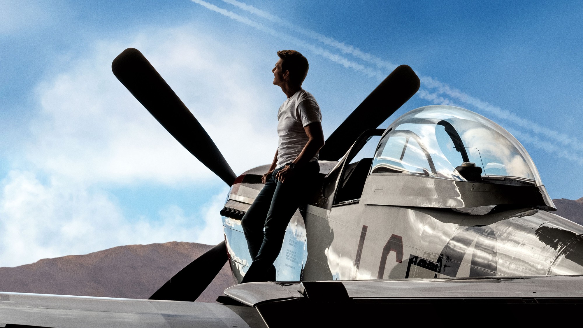 Tom Cruise de pie sobre el ala de un avión de hélice plateado, con nubes y montañas al fondo, en Top Gun: Maverick