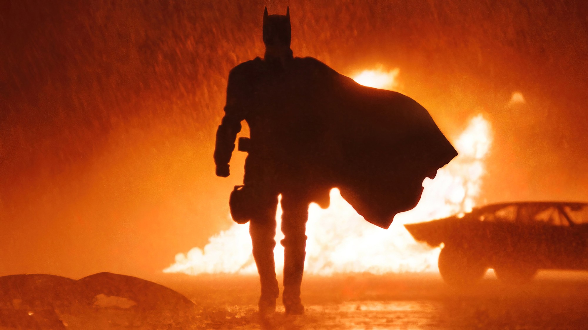 Silueta de Batman caminando bajo la lluvia frente a un incendio, con el Batimóvil detenido a un costado