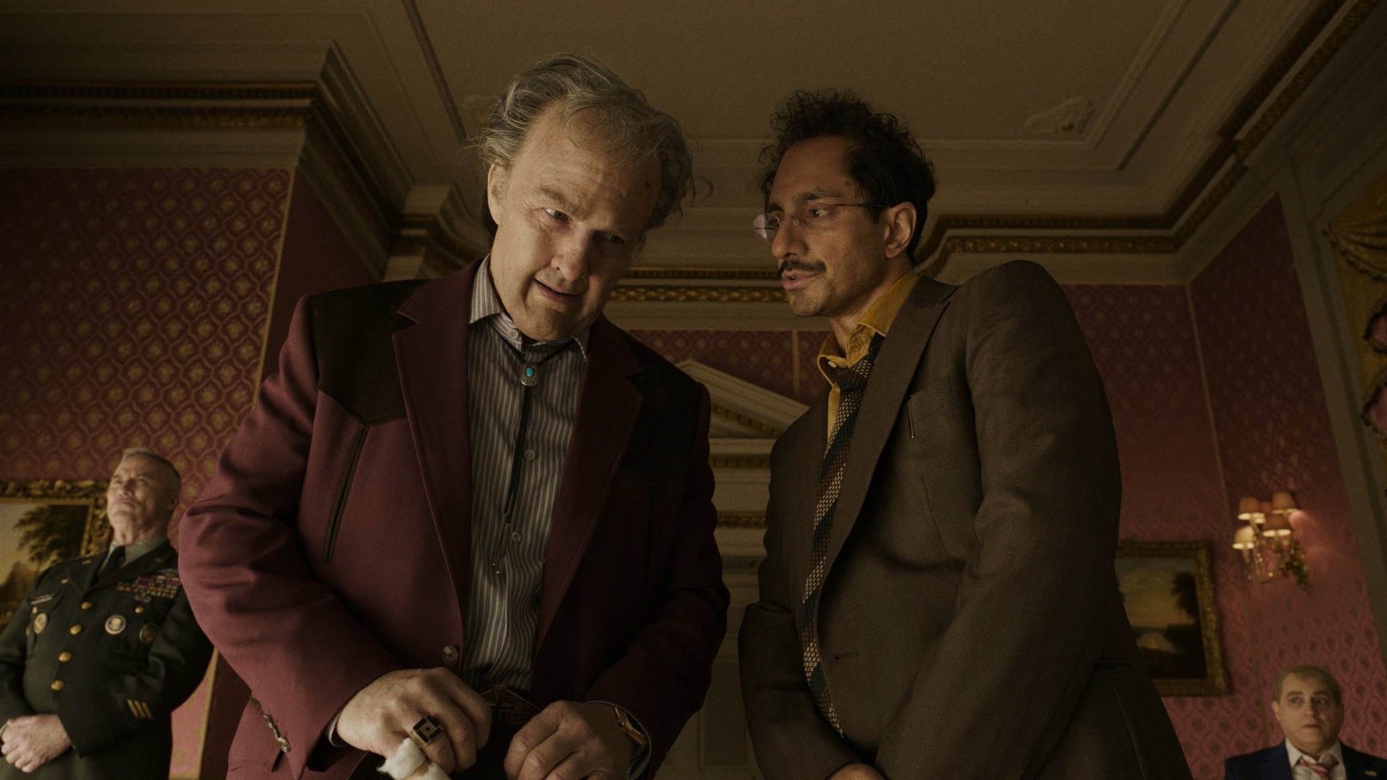 Tom Cruise como el magnate Digger Rockwell, con saco color vino, conversa con Riz Ahmed en un salón de paredes rojas