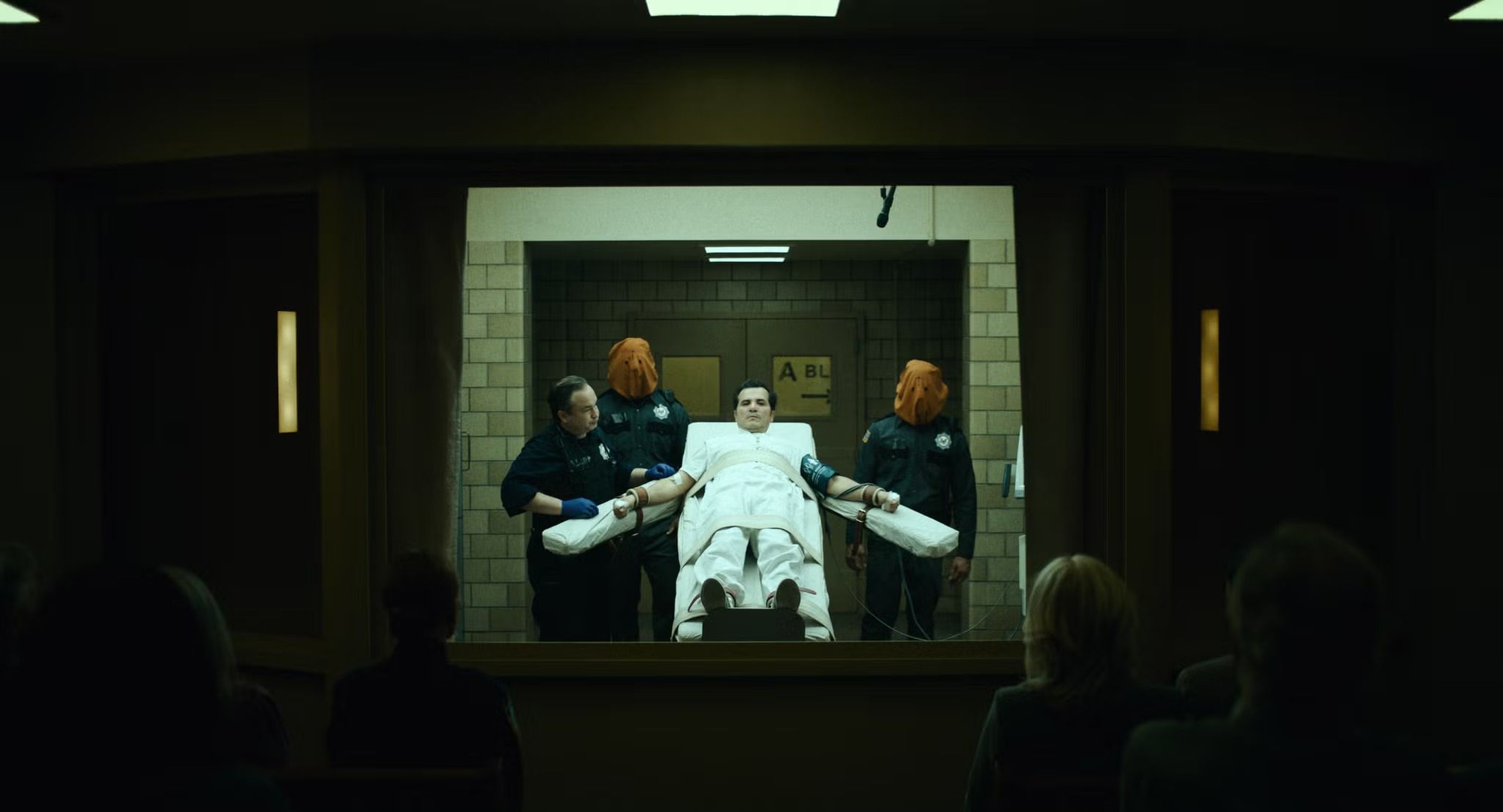 John Leguizamo como Father Sunday, vestido de blanco y atado a una camilla entre dos guardias con capuchas naranjas, observado por testigos tras un vidrio en The Exorcist: Martyrs