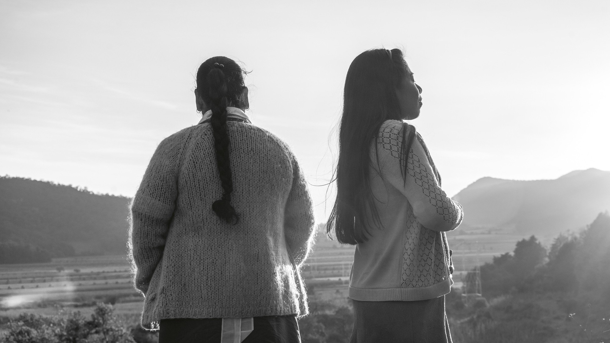 Dos mujeres de espaldas contemplan un valle con montañas al atardecer en una escena en blanco y negro de Roma