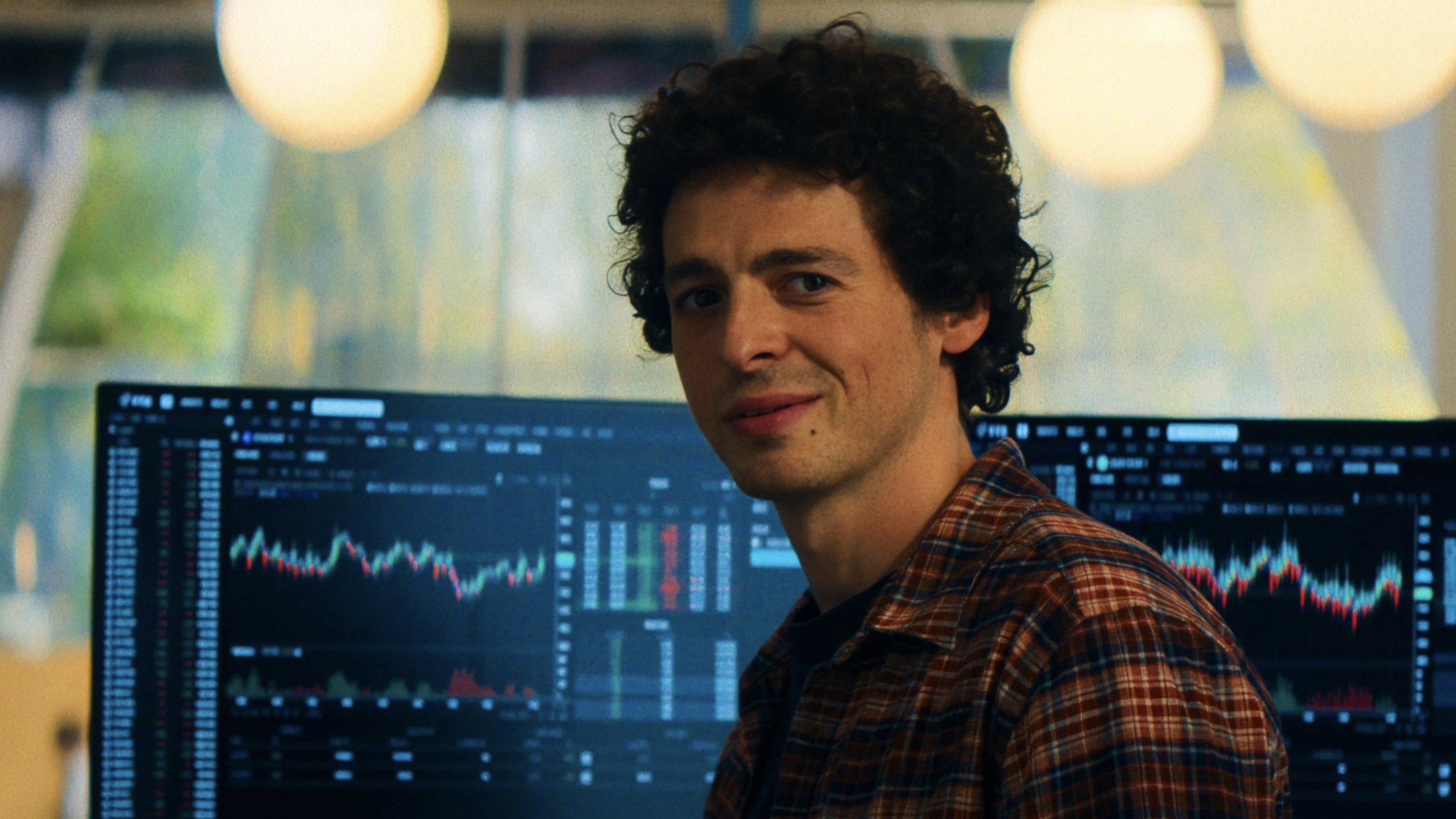 Anthony Boyle como Sam Bankman-Fried sonríe frente a varias pantallas con gráficos financieros en The Altruists