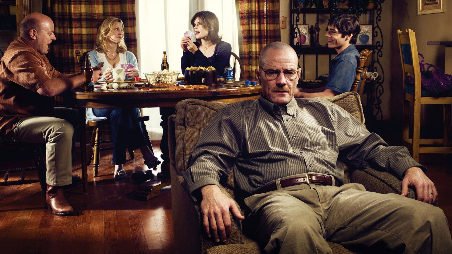 RJ Mitte sonríe al fondo mientras Anna Gunn, Betsy Brandt y Dean Norris juegan cartas y Bryan Cranston posa en un sillón en una imagen promocional de Breaking Bad