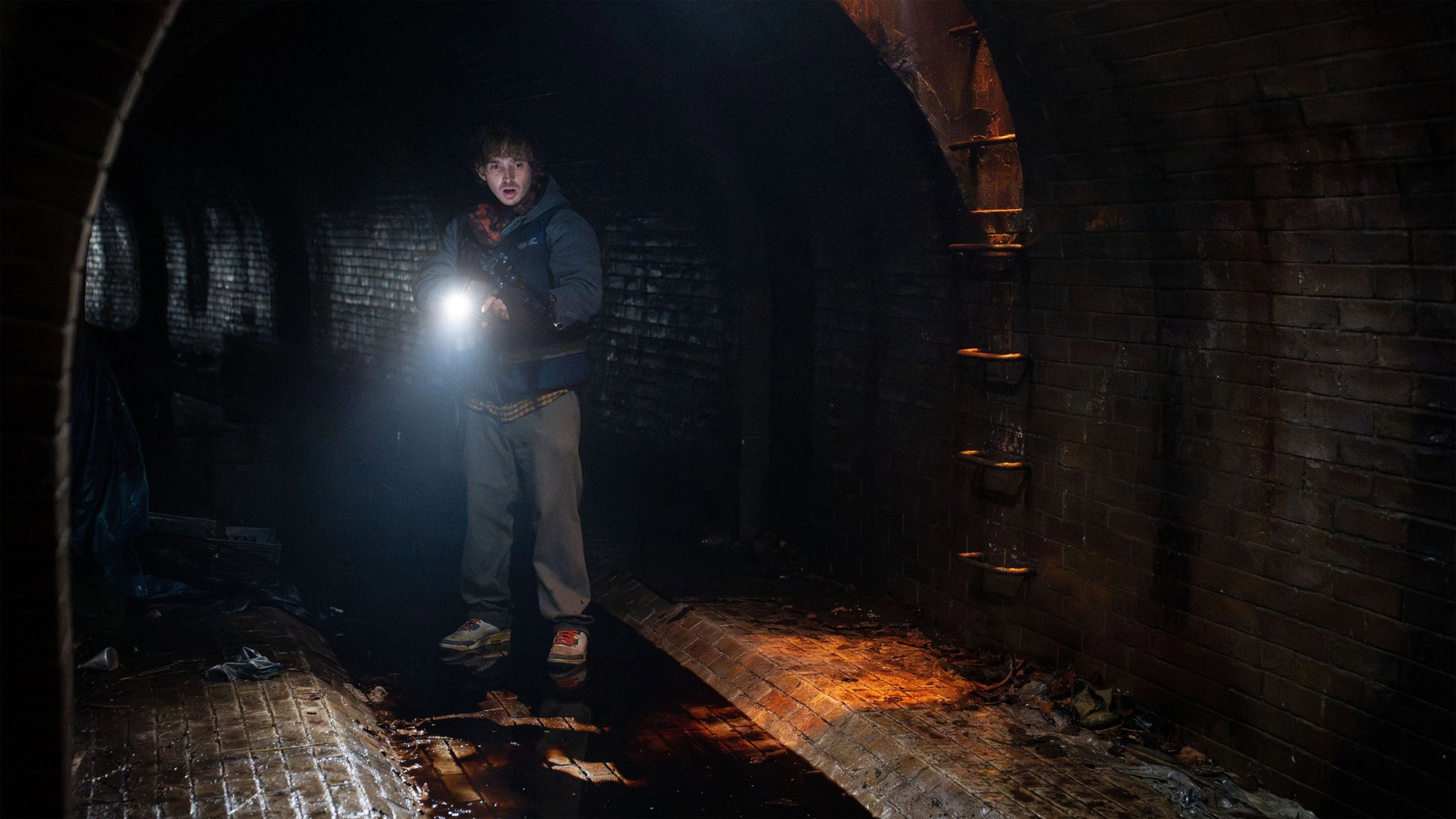 Bryan avanza con una linterna por un túnel de ladrillo oscuro y húmedo en Resident Evil: Noche Cero