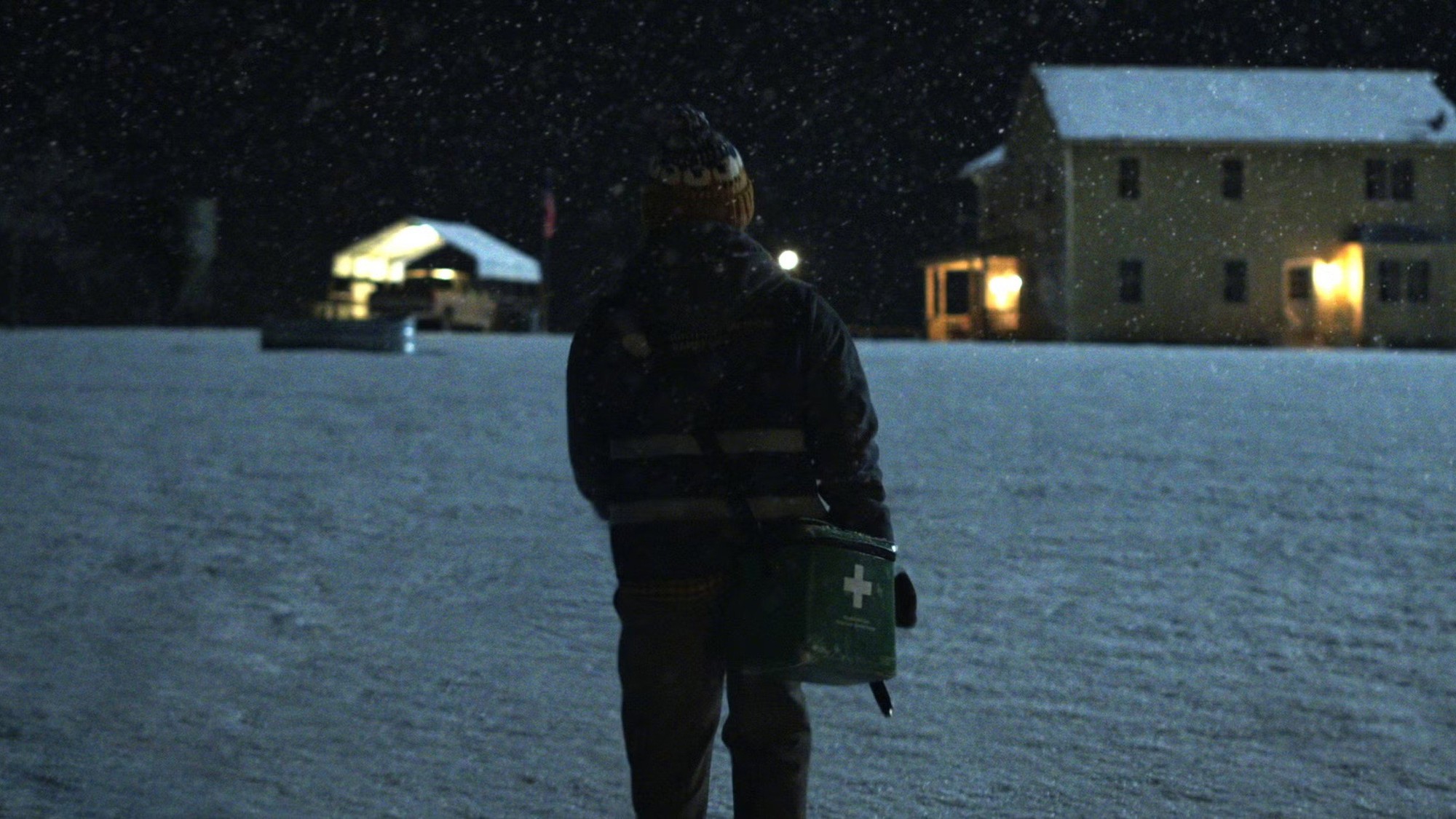 Un mensajero médico camina de espaldas bajo la nevada hacia una casa iluminada en medio de un campo cubierto de nieve