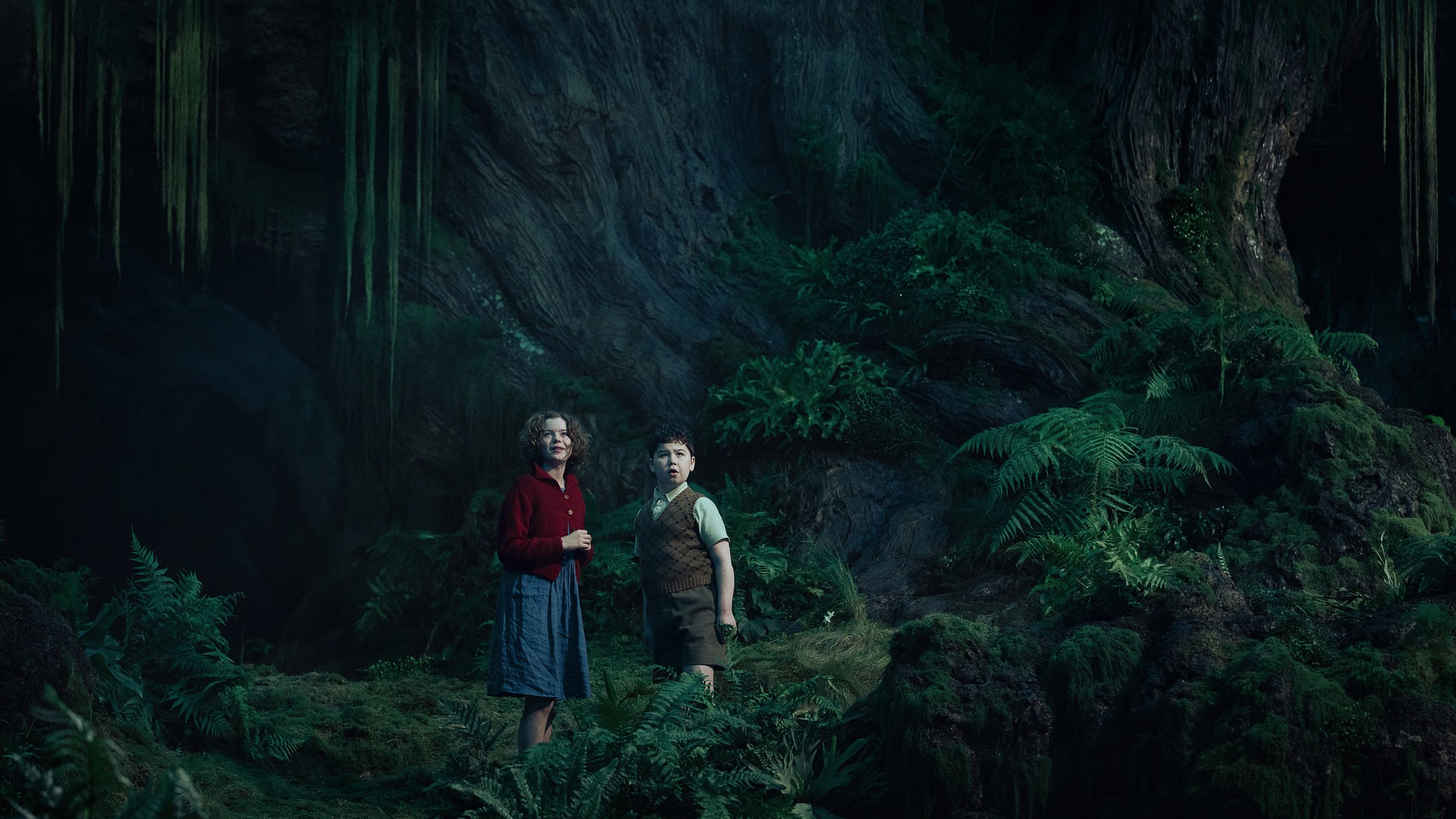 Polly y Digory observan asombrados un bosque oscuro y frondoso, cubierto de helechos y musgo, en Narnia: El sobrino del mago