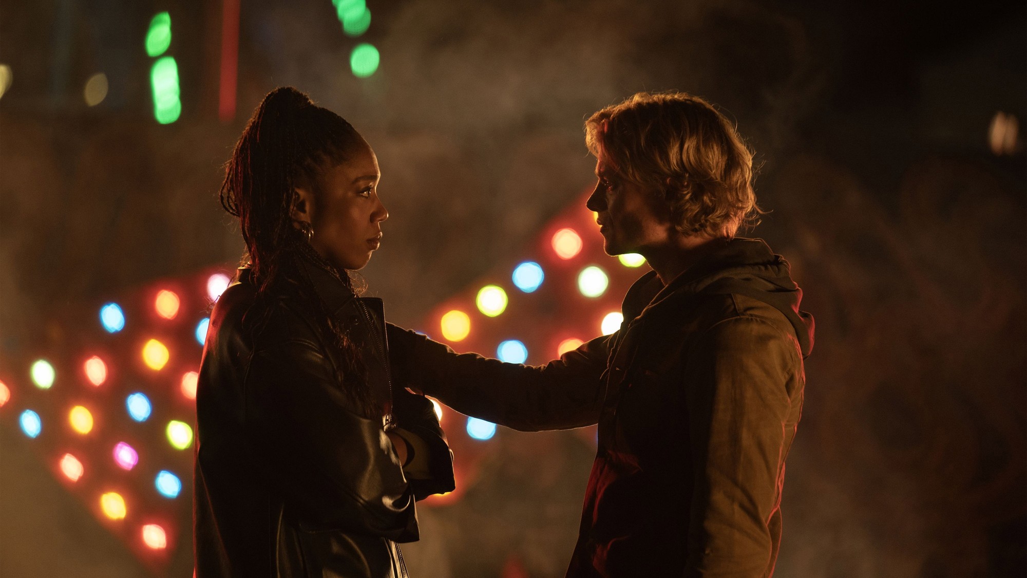 Naomi Ackie y Tom Rhys Harries se miran de frente en una calle nocturna con luces de colores desenfocadas en Clayface