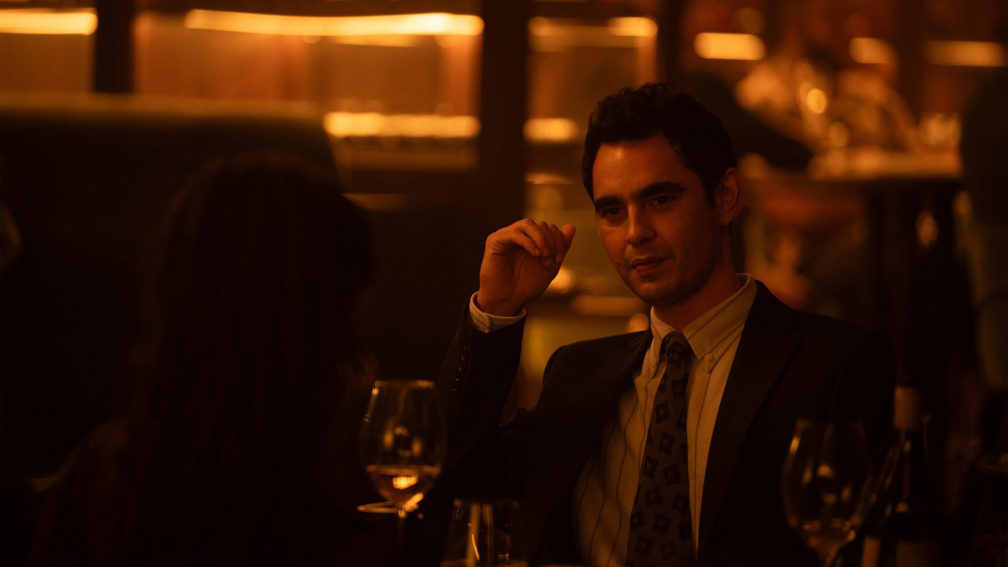 Max Minghella, de traje y corbata, conversa en la mesa de un restaurante con luz cálida y tenue en Clayface