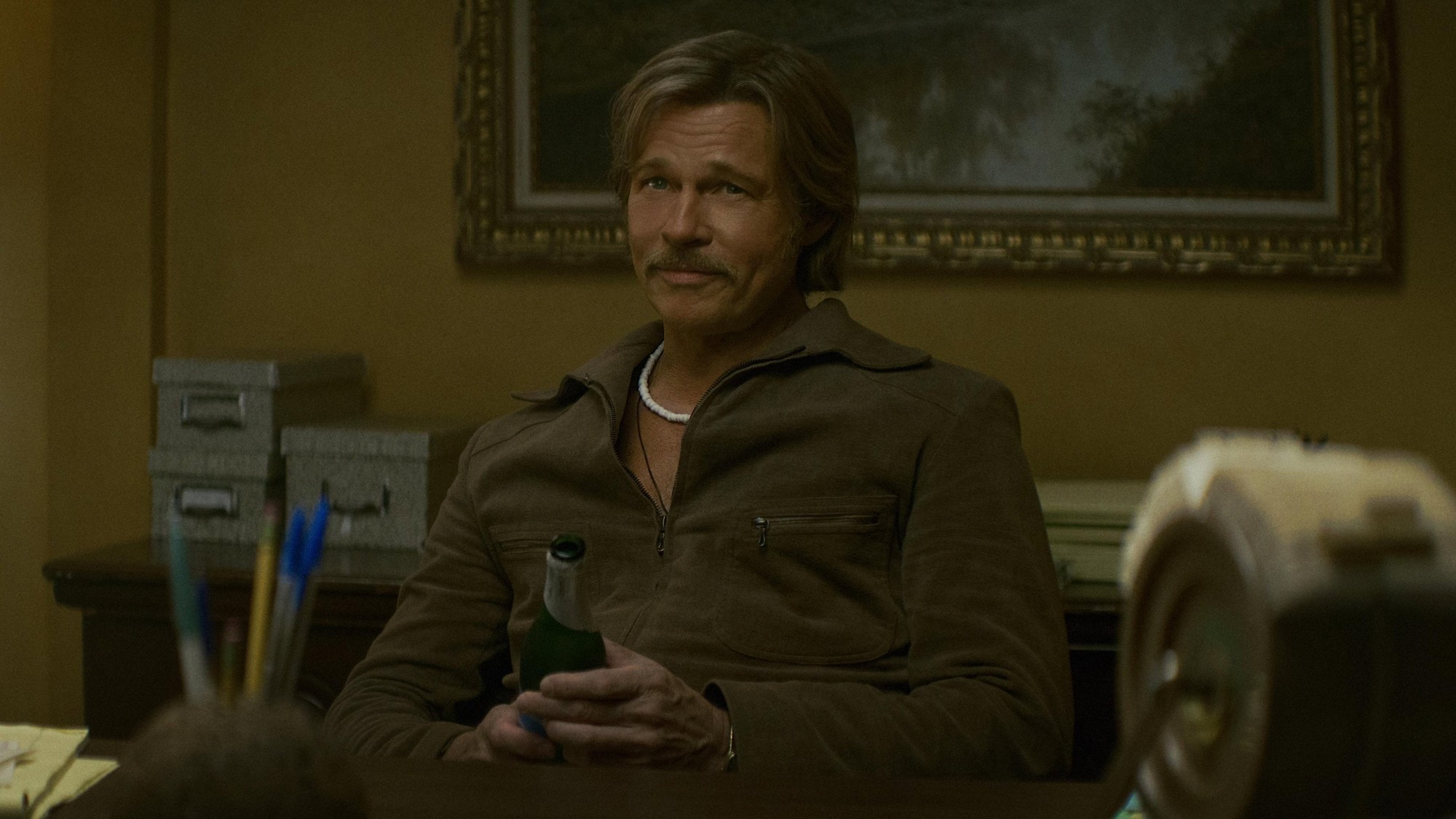 Brad Pitt como Cliff Booth, con chaqueta café y una cerveza en la mano, sonríe sentado frente a un escritorio