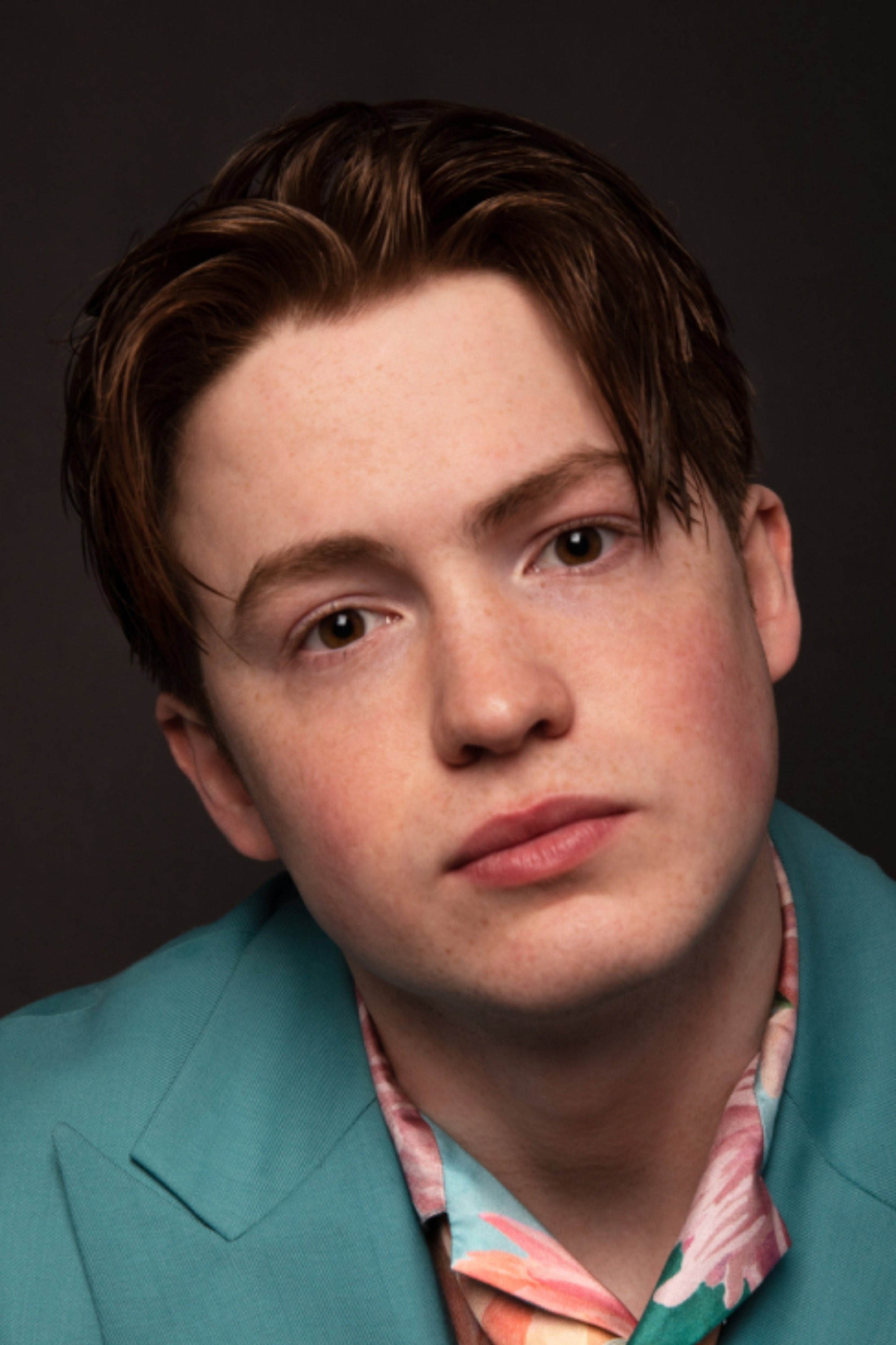 Retrato del actor Kit Connor con saco verde azulado y camisa estampada sobre fondo oscuro
