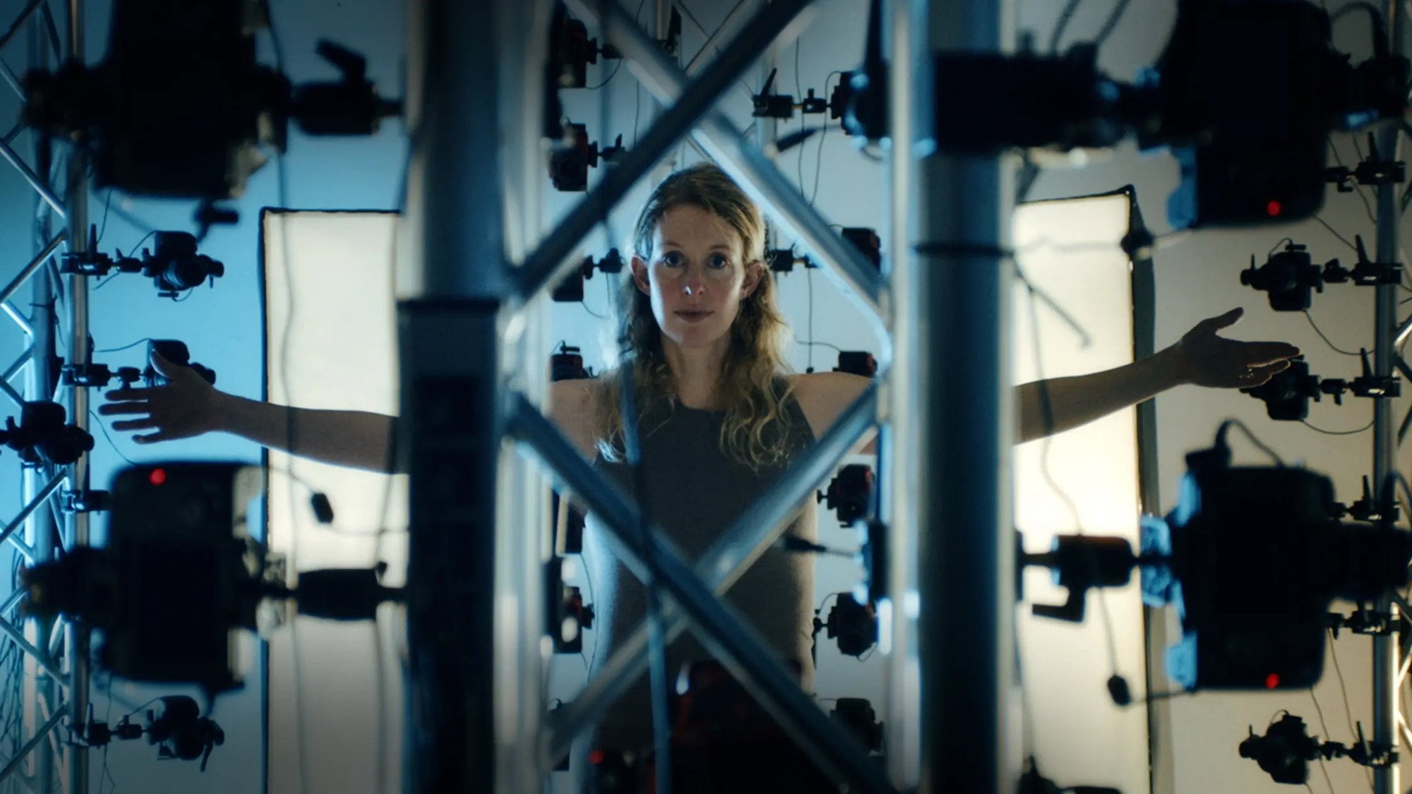 Elizabeth Holmes con los brazos extendidos entre estructuras metálicas y decenas de cámaras en el documental You Can See Everything