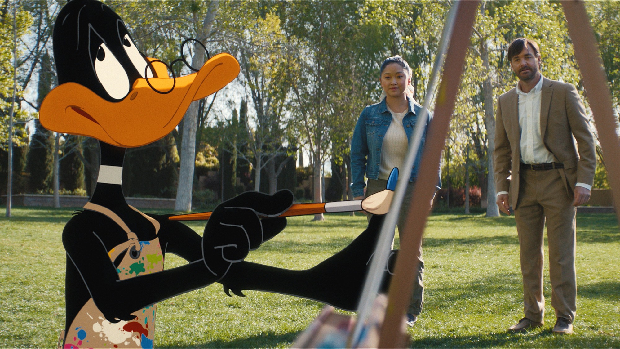 El Pato Lucas con delantal de pintor y pincel en mano frente a Lana Condor y Will Forte en un parque, escena de Coyote vs. Acme