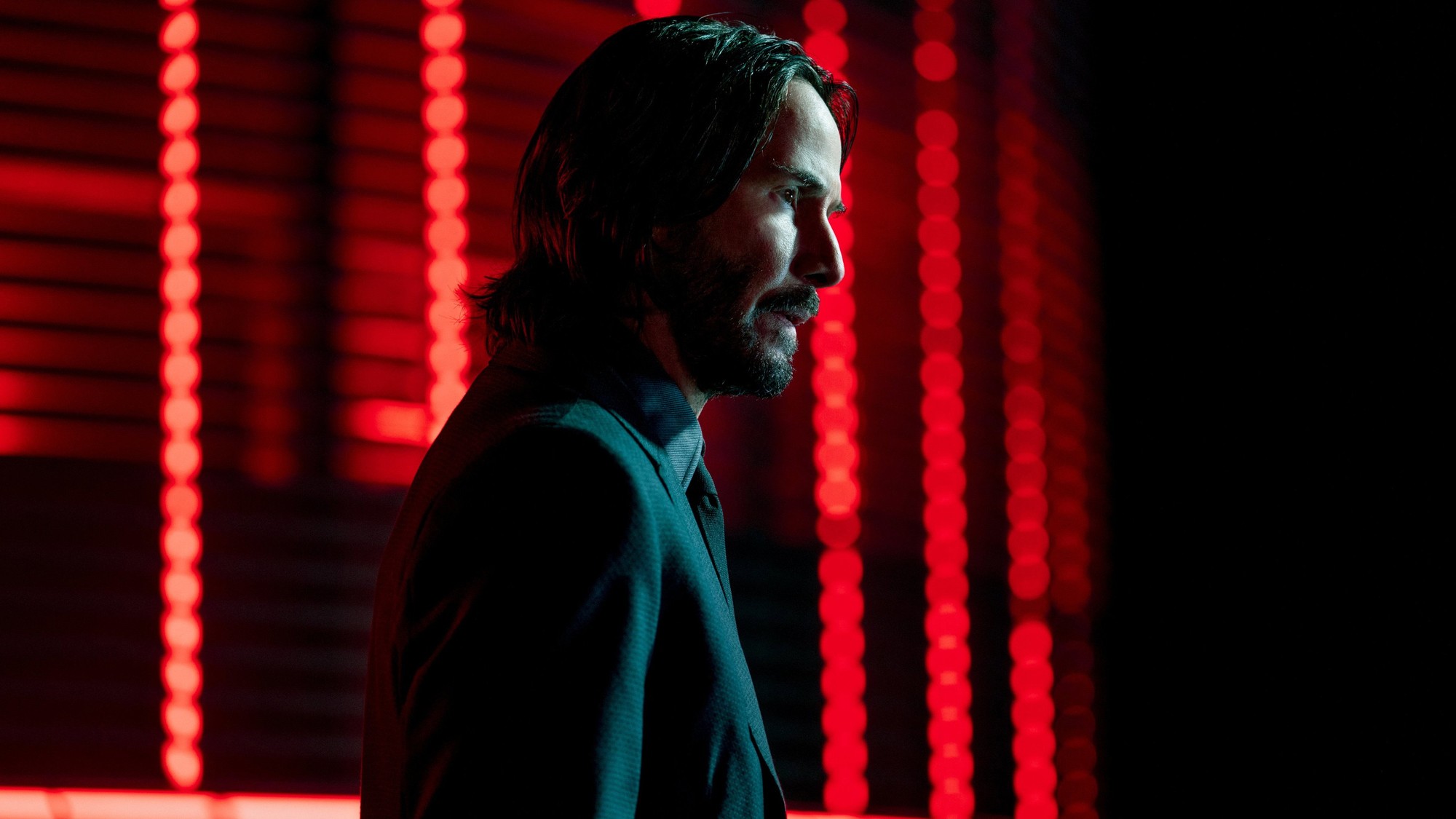 Keanu Reeves de perfil, con barba y traje oscuro, frente a un fondo de luces rojas en John Wick: Capítulo 4
