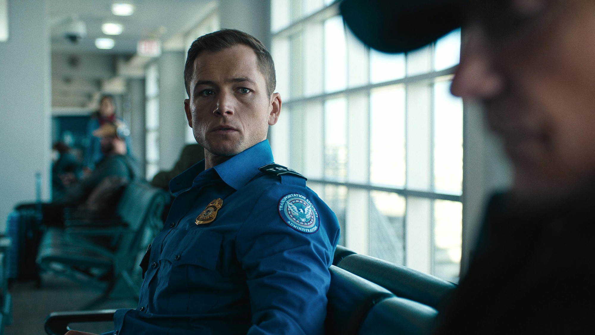 Taron Egerton con uniforme de agente de seguridad, sentado en la sala de espera de un aeropuerto en Carry-On