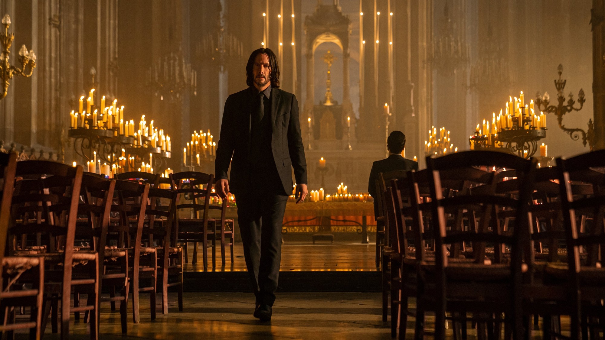 Keanu Reeves camina con traje negro por el pasillo central de una iglesia iluminada con velas en John Wick: Capítulo 4
