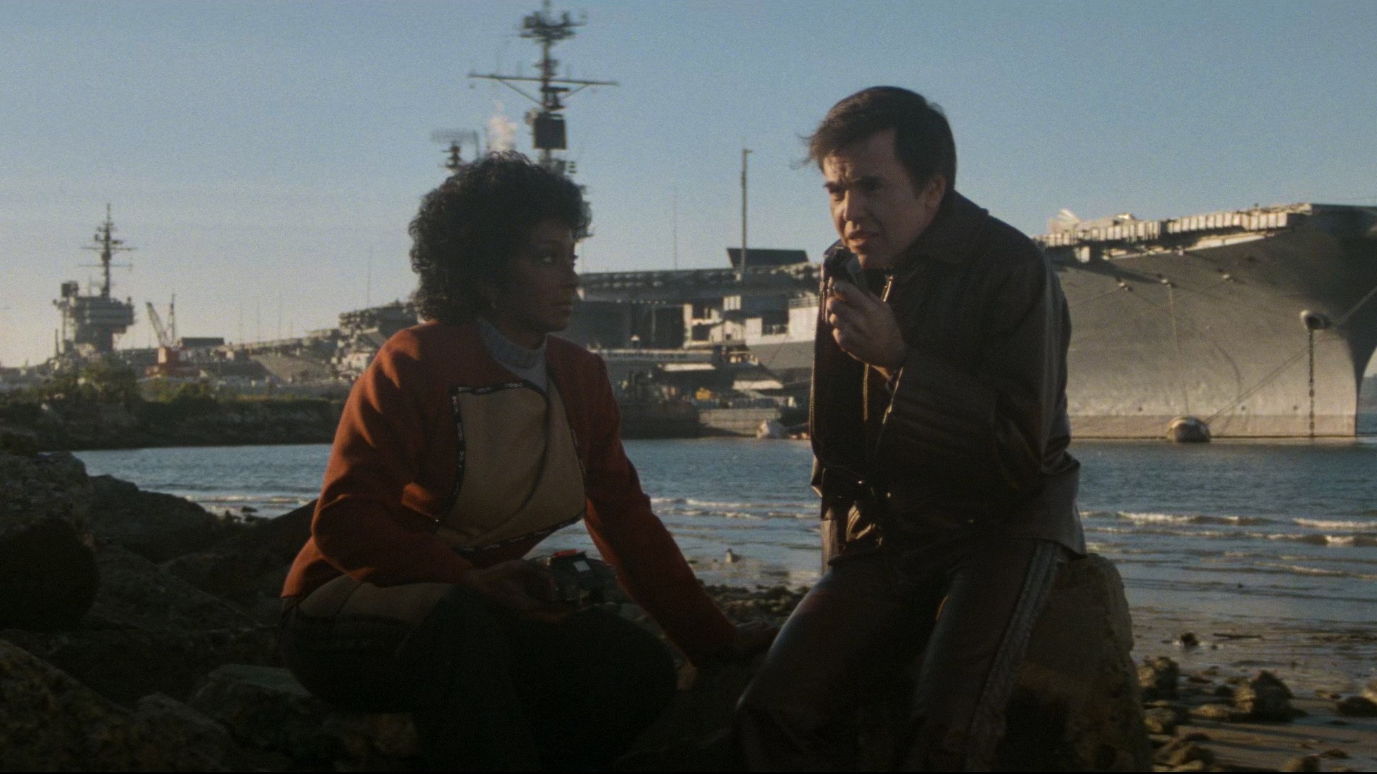 Walter Koenig como Chekov habla por un comunicador junto a Nichelle Nichols como Uhura, frente a un portaaviones en Star Trek IV: The Voyage Home