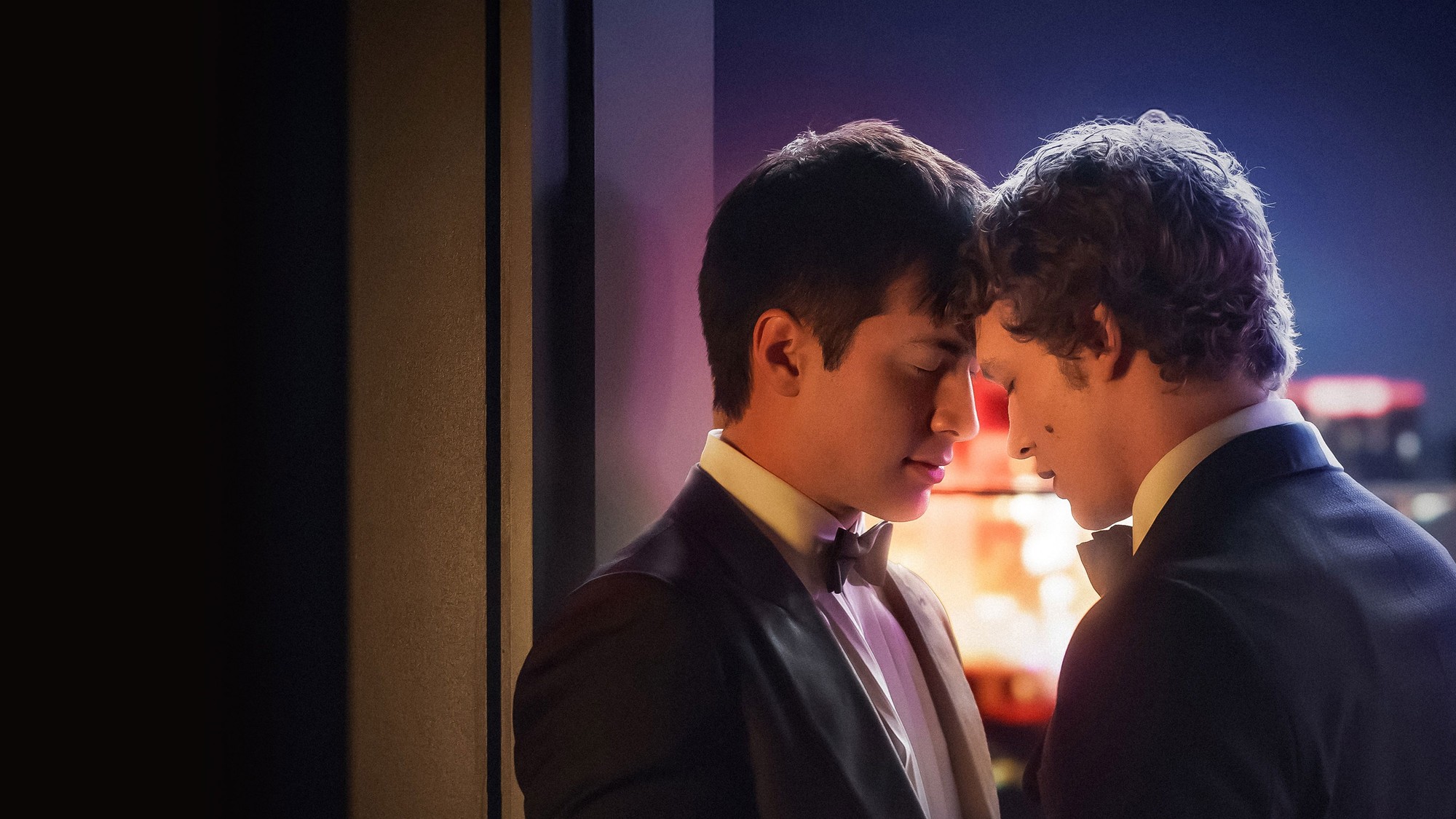 Connor Storrie y Hudson Williams, vestidos de esmoquin, juntan sus frentes frente a una ventana con luces de ciudad en Heated Rivalry
