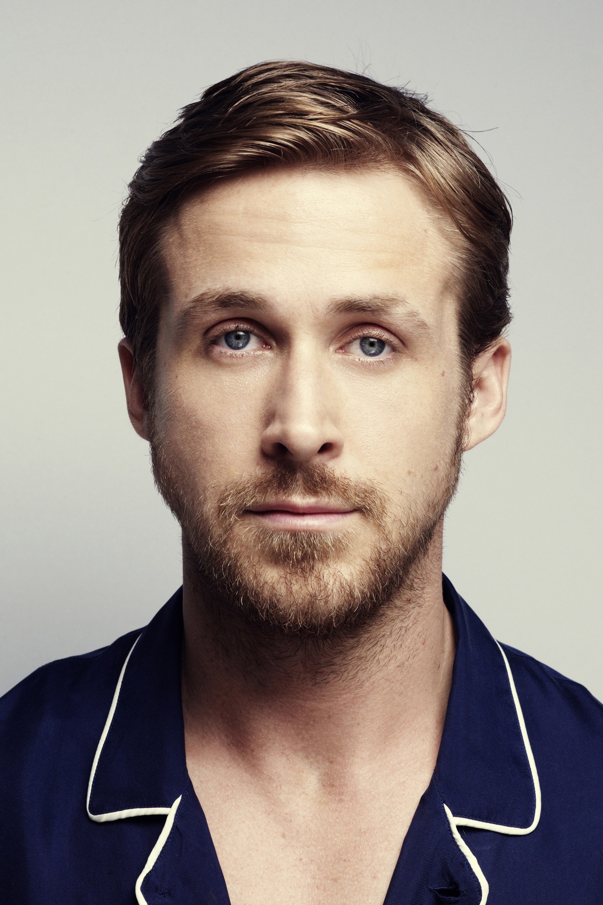 Retrato de estudio de Ryan Gosling con barba corta y camisa azul oscuro sobre un fondo gris claro