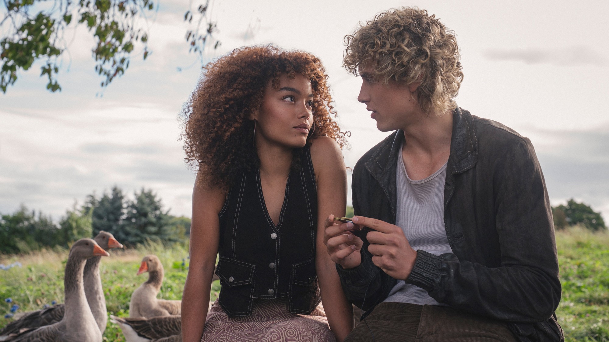 Whitney Peak como Lenore Dove y Joseph Zada como el joven Haymitch Abernathy conversan sentados en un prado junto a unos gansos en Amanecer en la cosecha