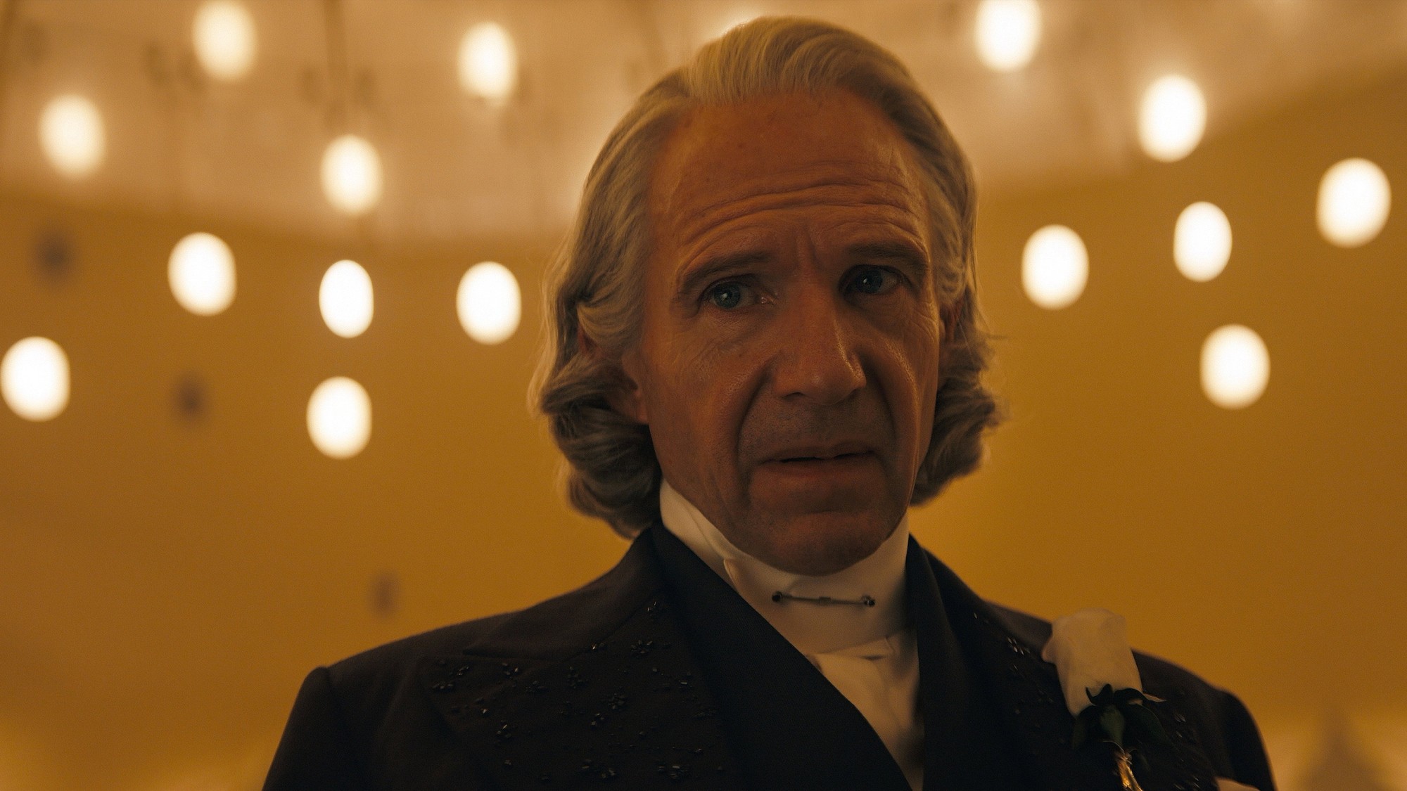 Ralph Fiennes como el presidente Snow, con el cabello gris largo y una rosa blanca en la solapa, bajo luces cálidas en Amanecer en la cosecha