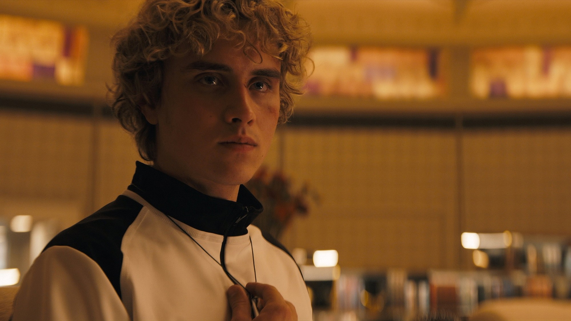 Joseph Zada como el joven Haymitch Abernathy, con el cabello rizado y una chaqueta blanca y negra, mira a cámara bajo una luz cálida en Amanecer en la cosecha