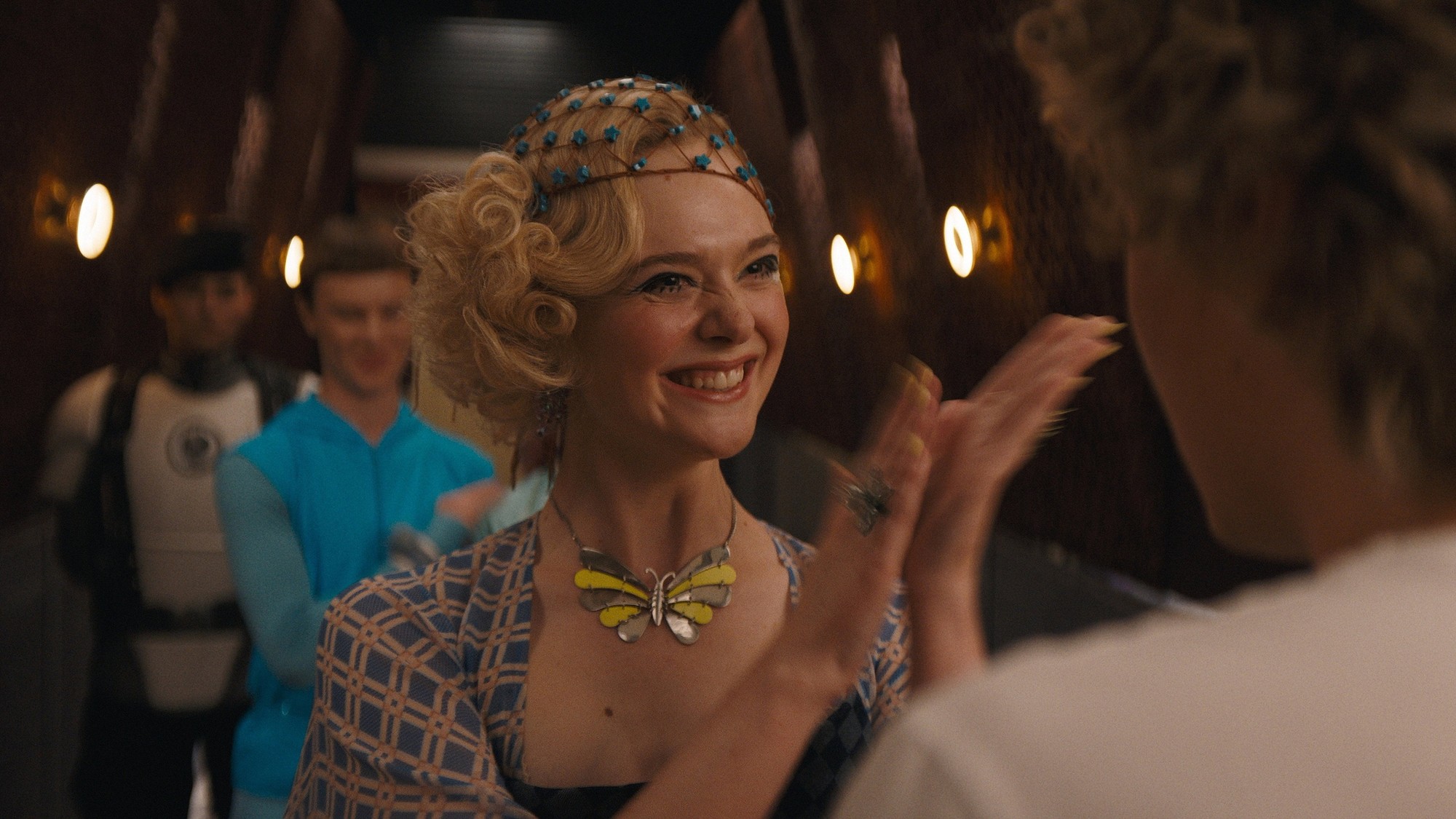 Elle Fanning como una joven Effie Trinket, con rizos rubios y un collar de mariposa, aplaude sonriente en un pasillo del Capitolio en Amanecer en la cosecha