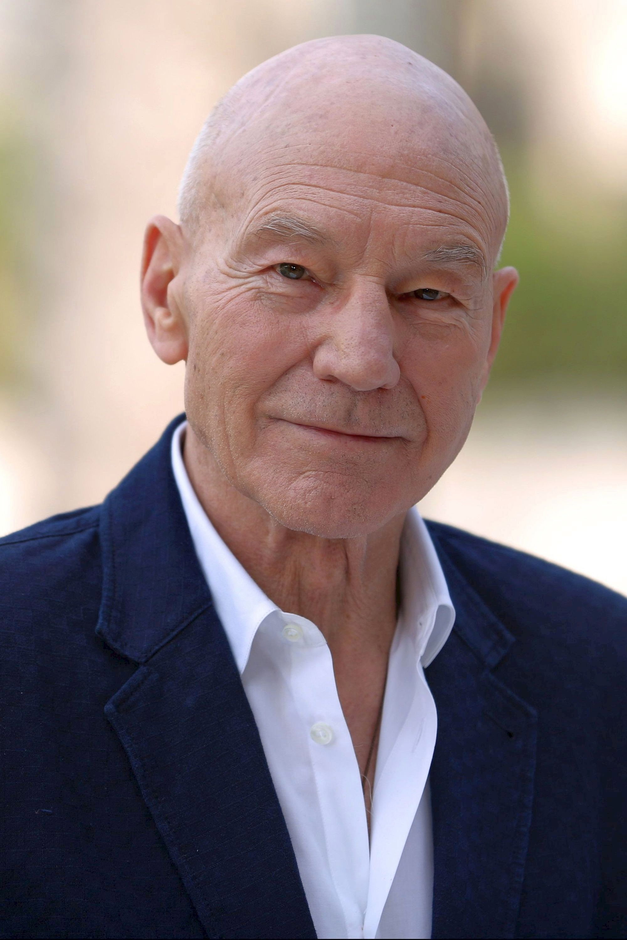 Retrato de Patrick Stewart sonriendo, con saco azul marino y camisa blanca, al aire libre